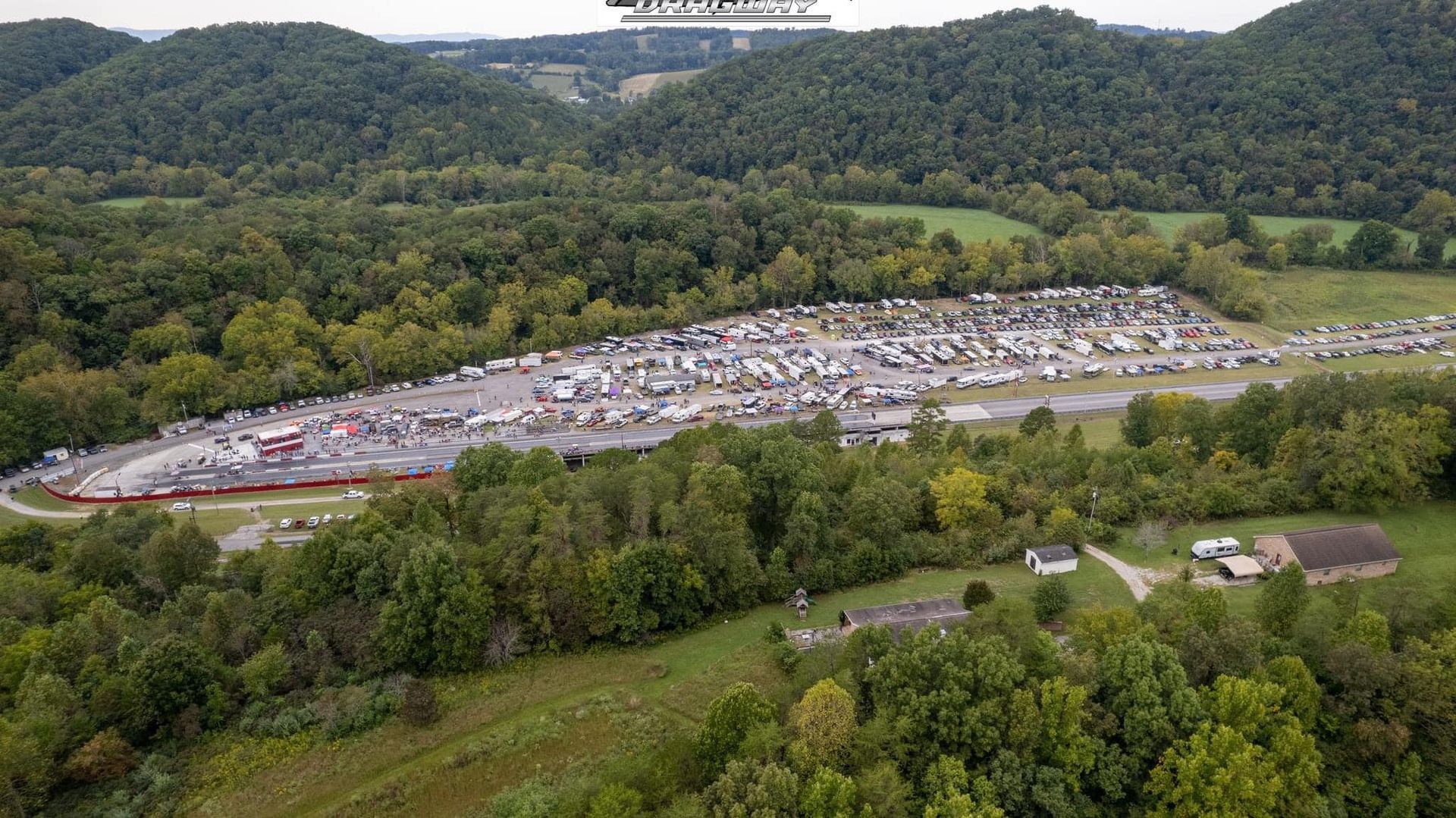 Aerial view of a large parking lot by a road, surrounded by dense green forest and hills.