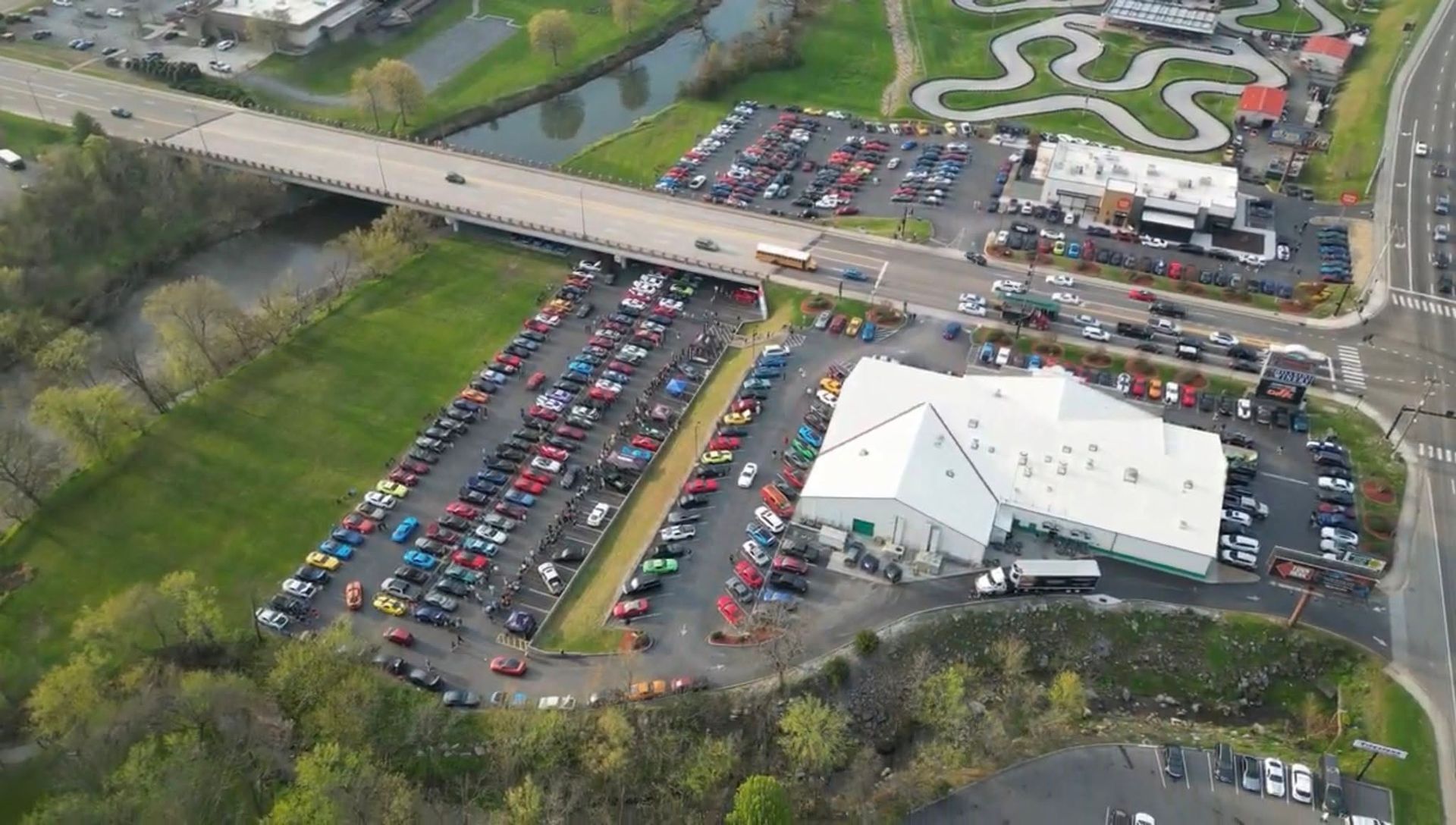 Aerial view of a large parking lot beside a white-roofed building and bridge, with many parked cars and green fields nearby
