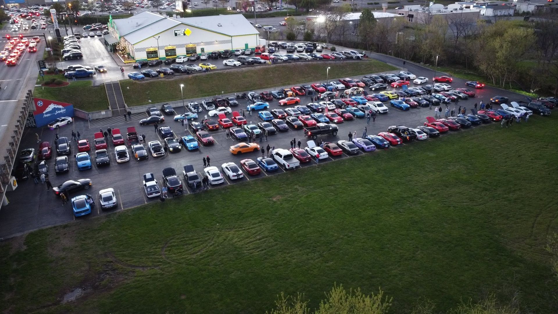 Aerial view of a large parking lot full of cars beside a white building and grassy field