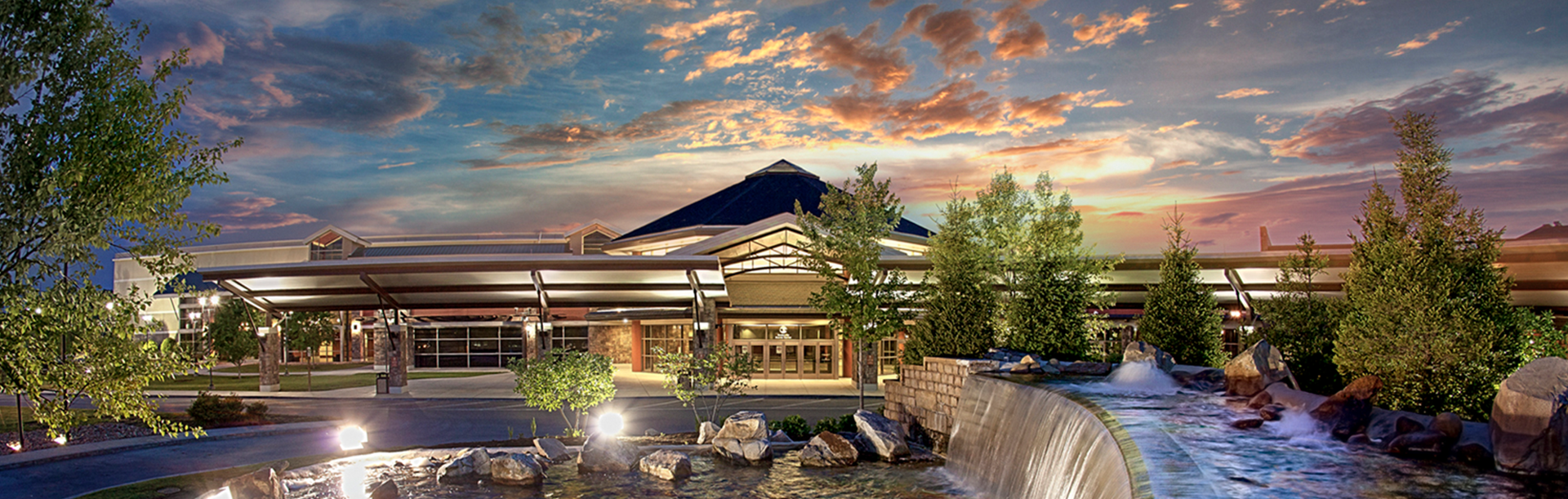 Modern lodge at sunset with mountains, trees, and a flowing stream in the foreground