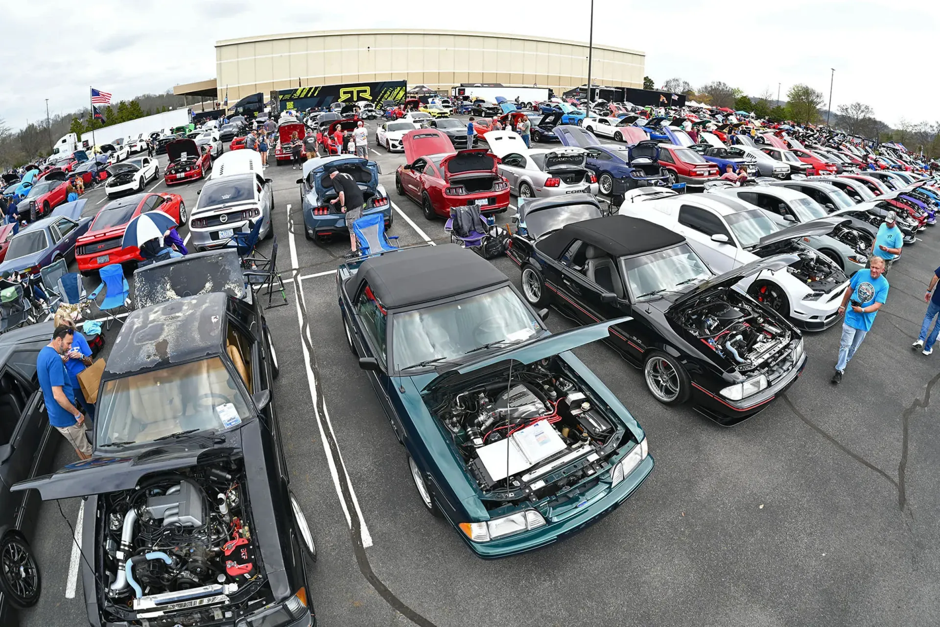 Crowded car show with rows of customized vehicles and people gathered outside a large building