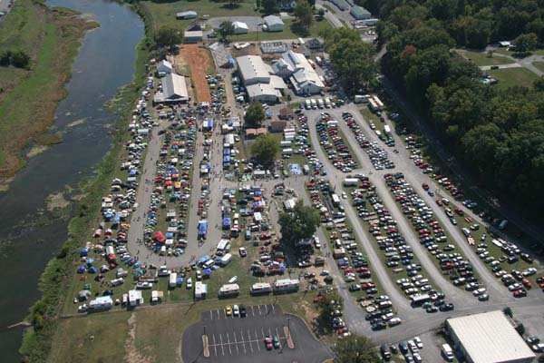 Aerial view of a crowded outdoor car show with rows of parked cars and tents beside a river.