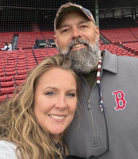 Smiling couple at a baseball stadium, with red seats and a Boston Red Sox logo on his jacket