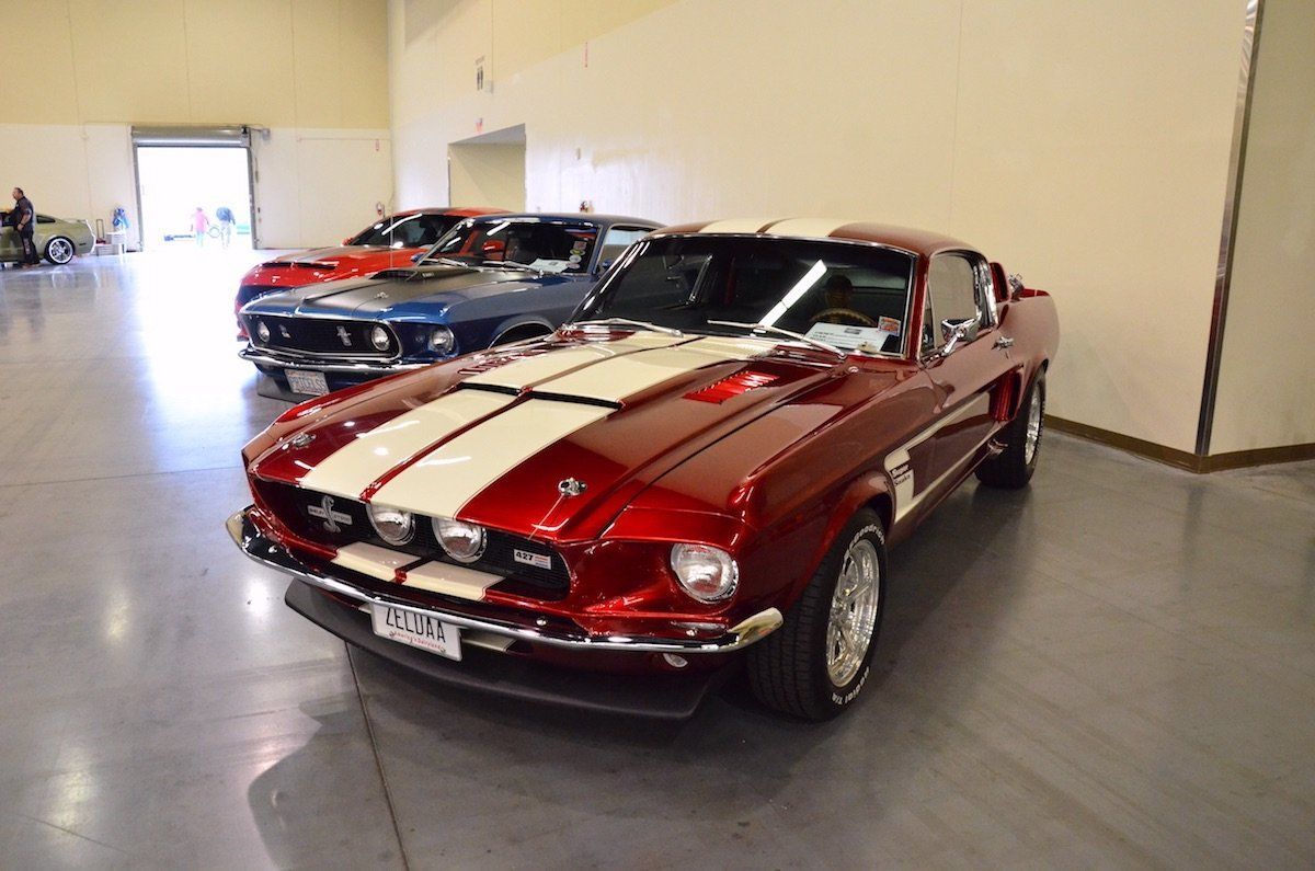 Red classic muscle car with white racing stripes parked indoors beside other cars.