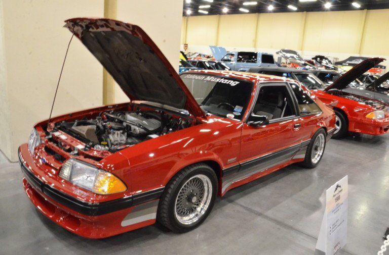 Red sports car with hood open at an indoor car show, surrounded by other classic cars