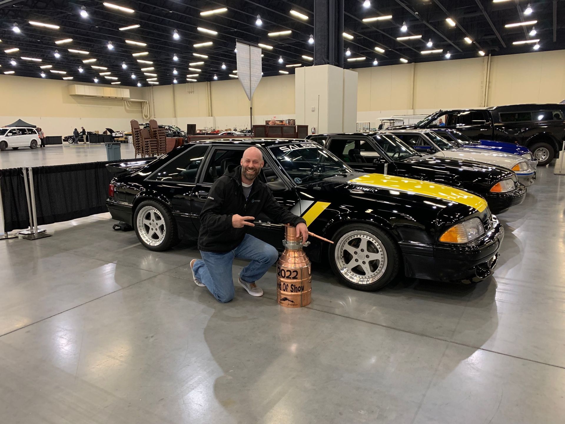 Man crouching beside a black sports car with yellow stripes at an indoor car show, holding a bottle