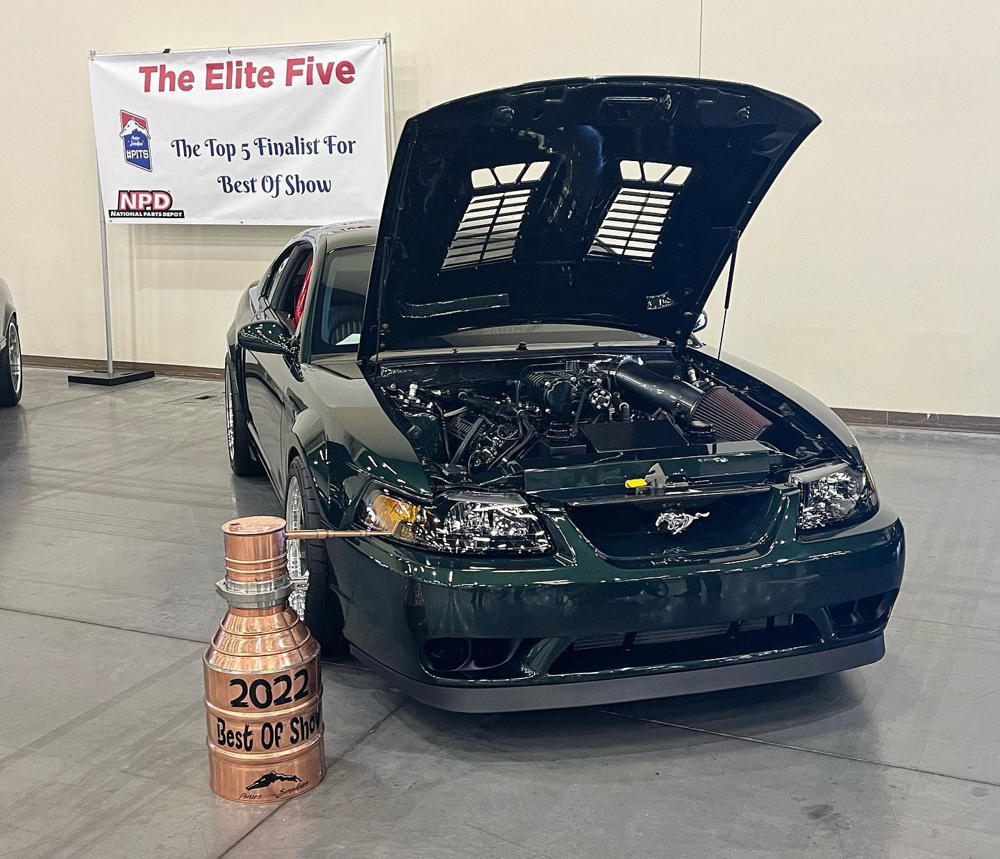 Dark green car with hood open on display, beside a copper trophy, at The Elite Five event.