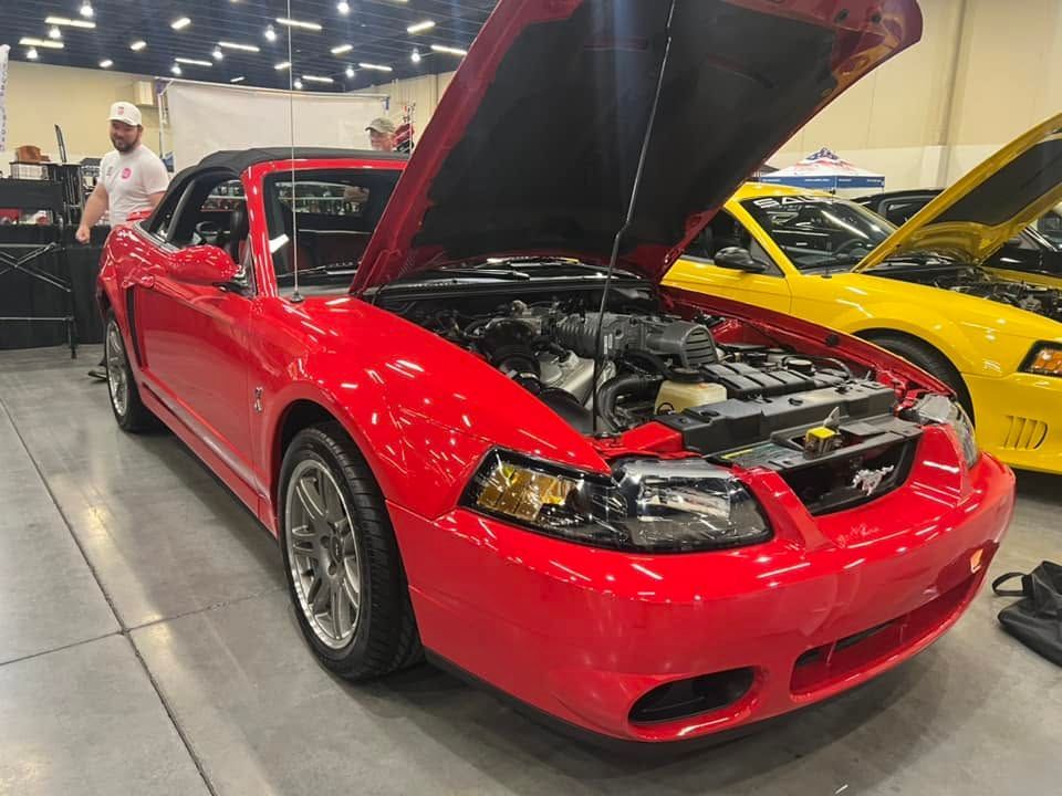 Red sports car with hood open at an indoor car show, with a yellow car beside it