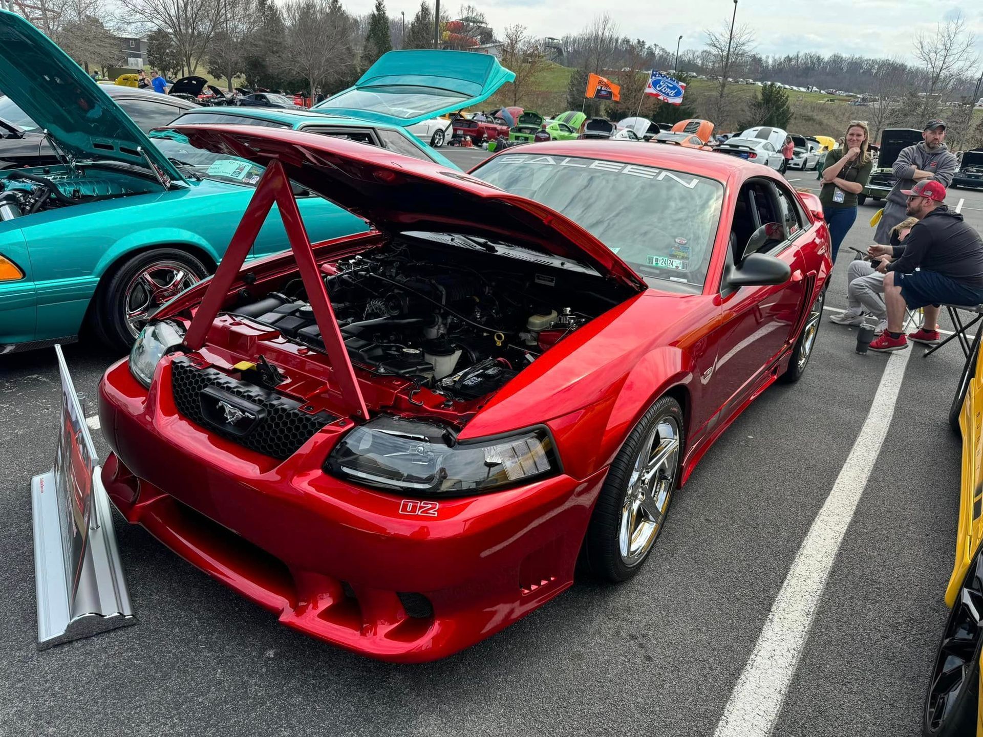 Red sports car with hood open at an outdoor car show, with a teal car beside it.