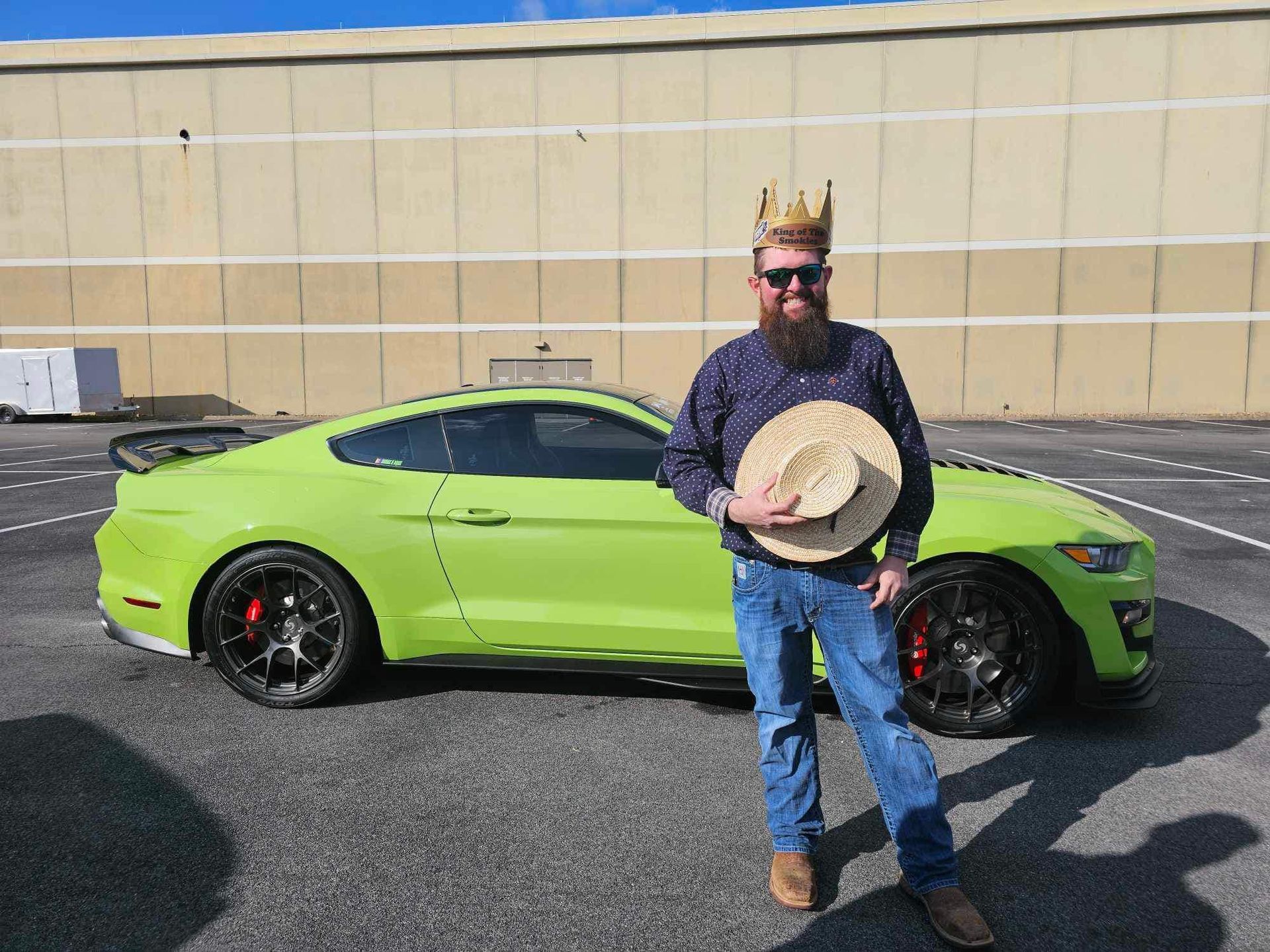 Man wearing a crown stands holding a hat in front of a bright green sports car outdoors.