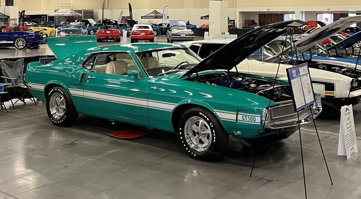 Turquoise classic convertible car on display indoors with hood open and informational sign nearby