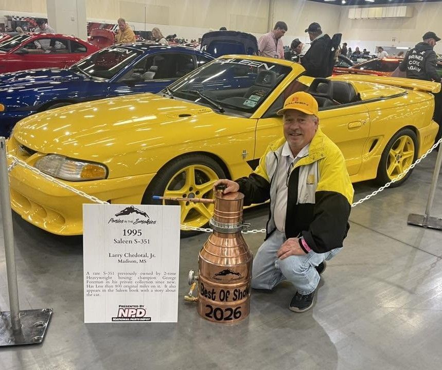Yellow sports car at an indoor show, with a man kneeling beside it and a copper trophy in front.