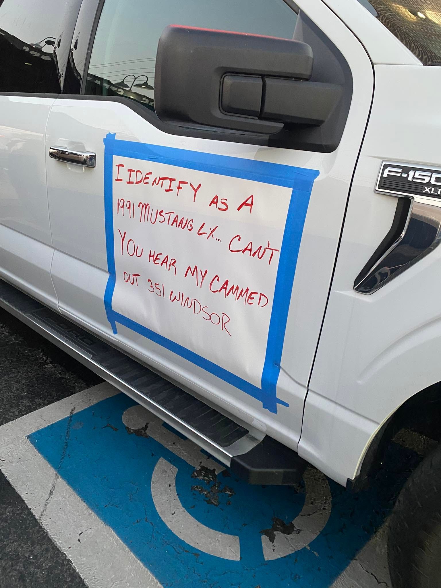 White pickup truck parked in an accessible space with a handwritten sign taped to the door.
