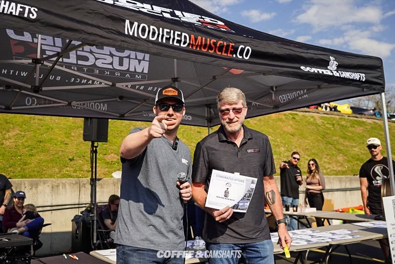 Two men holding an award plaque under a race event tent outdoors.