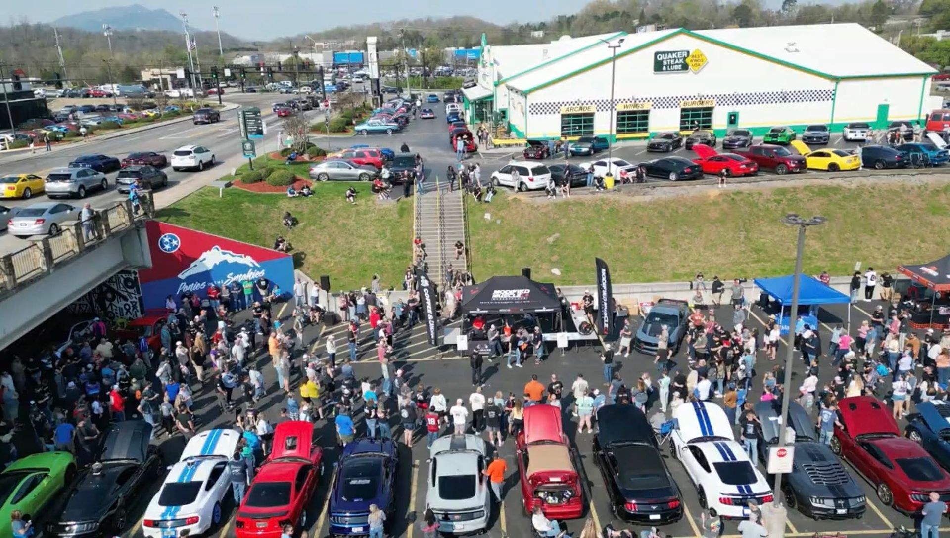 Crowded outdoor car show with rows of parked vehicles and people gathered beside a large white building