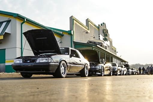 Classic cars lined up at a drive-in, with one black car’s hood open in the foreground.