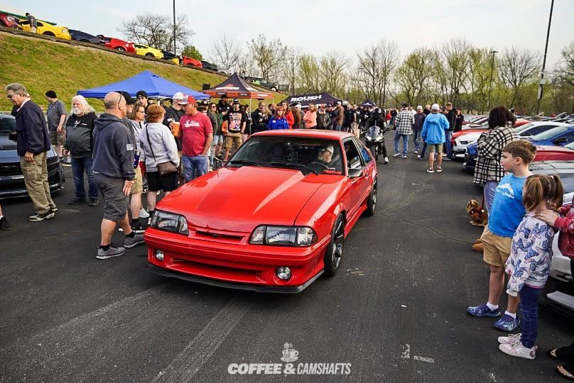 Red sports car at a crowded outdoor car show, with spectators lining both sides of the pavement.
