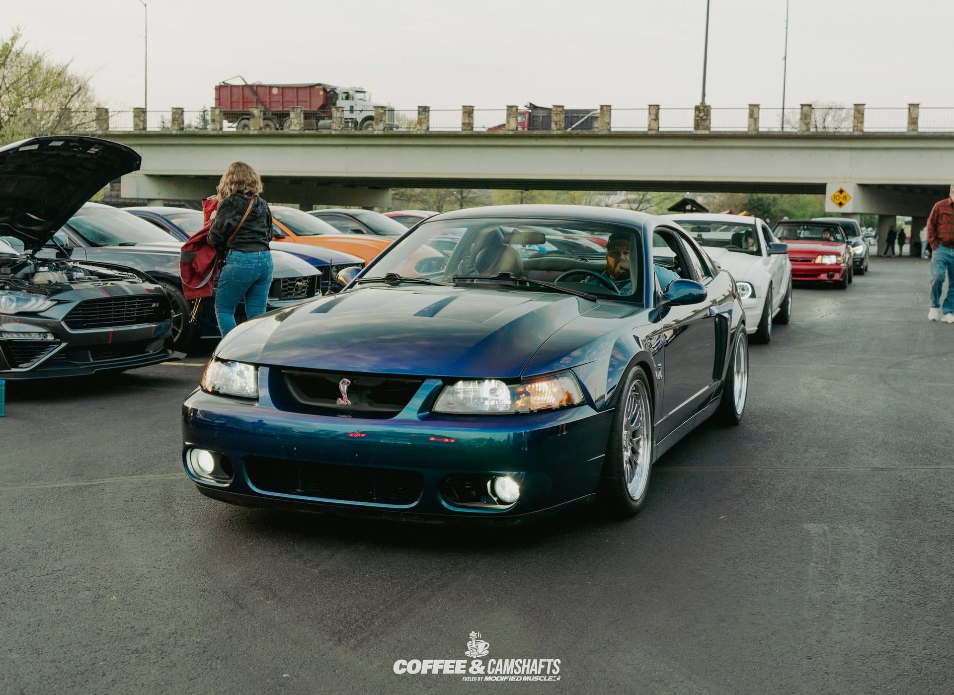 Blue Ford Mustang leading a line of cars at an outdoor meet, with one hood open and a person standing nearby
