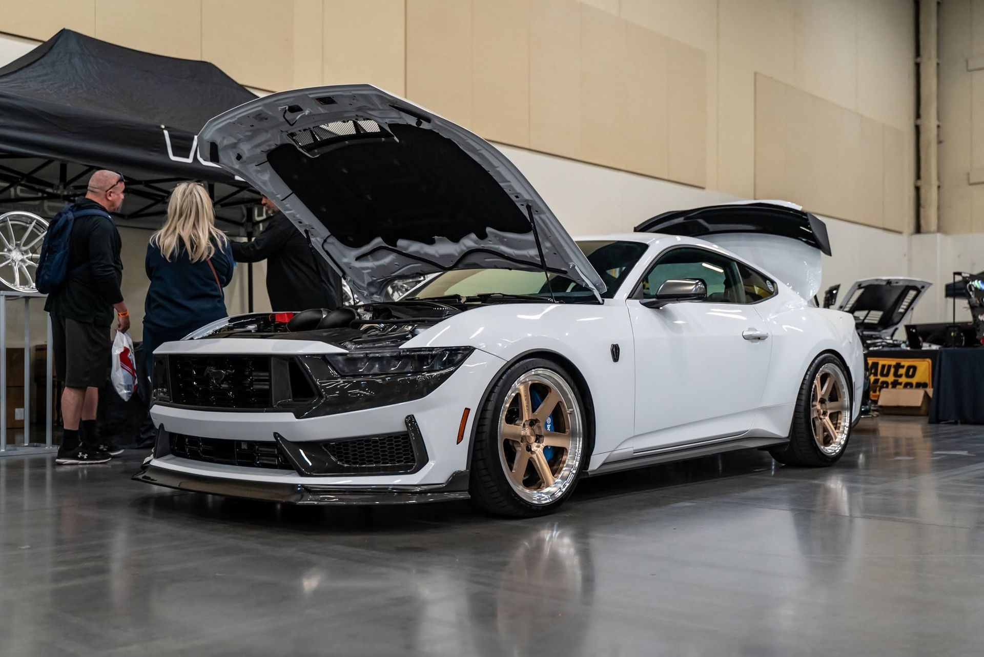White sports car with hood open at an indoor auto show, surrounded by people and other vehicles