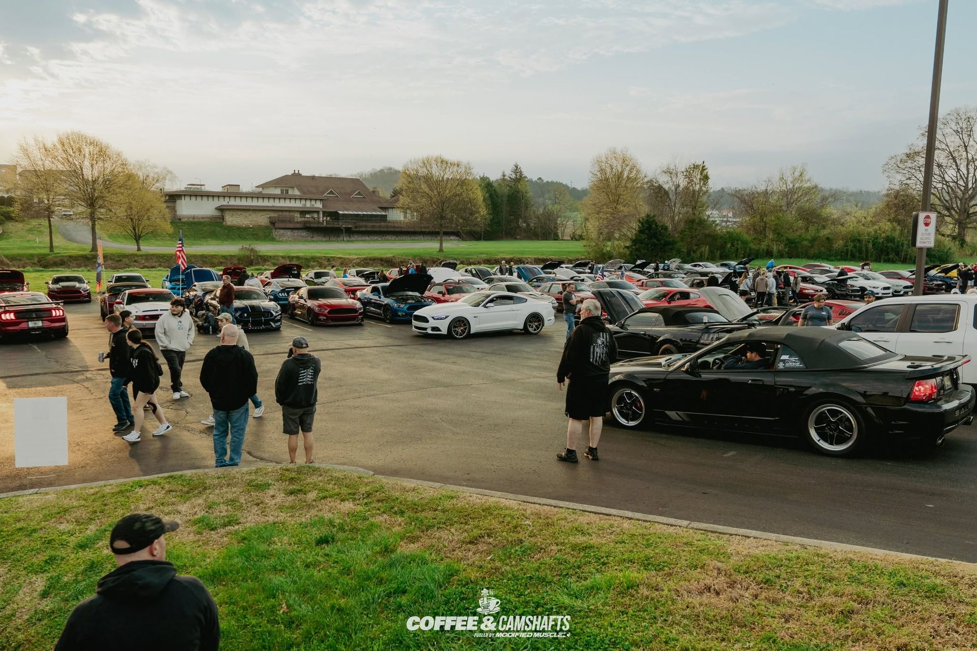 Parking lot with many cars and people gathered near a grassy field under cloudy skies