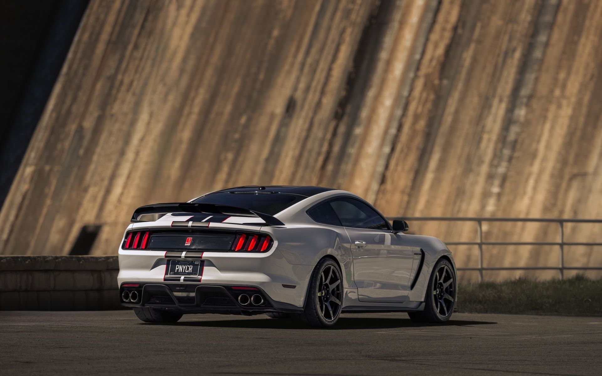 White sports car on a curved road at dusk, viewed from the rear against a wooden barrier.