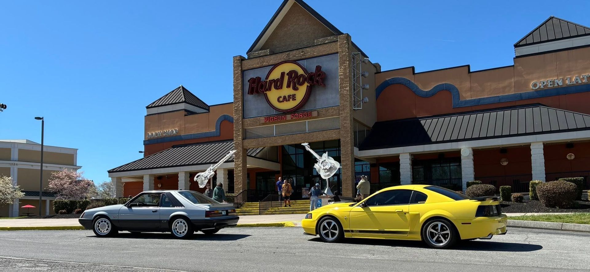 Hard Rock Cafe storefront with a black sedan and yellow sports car parked outside under a blue sky