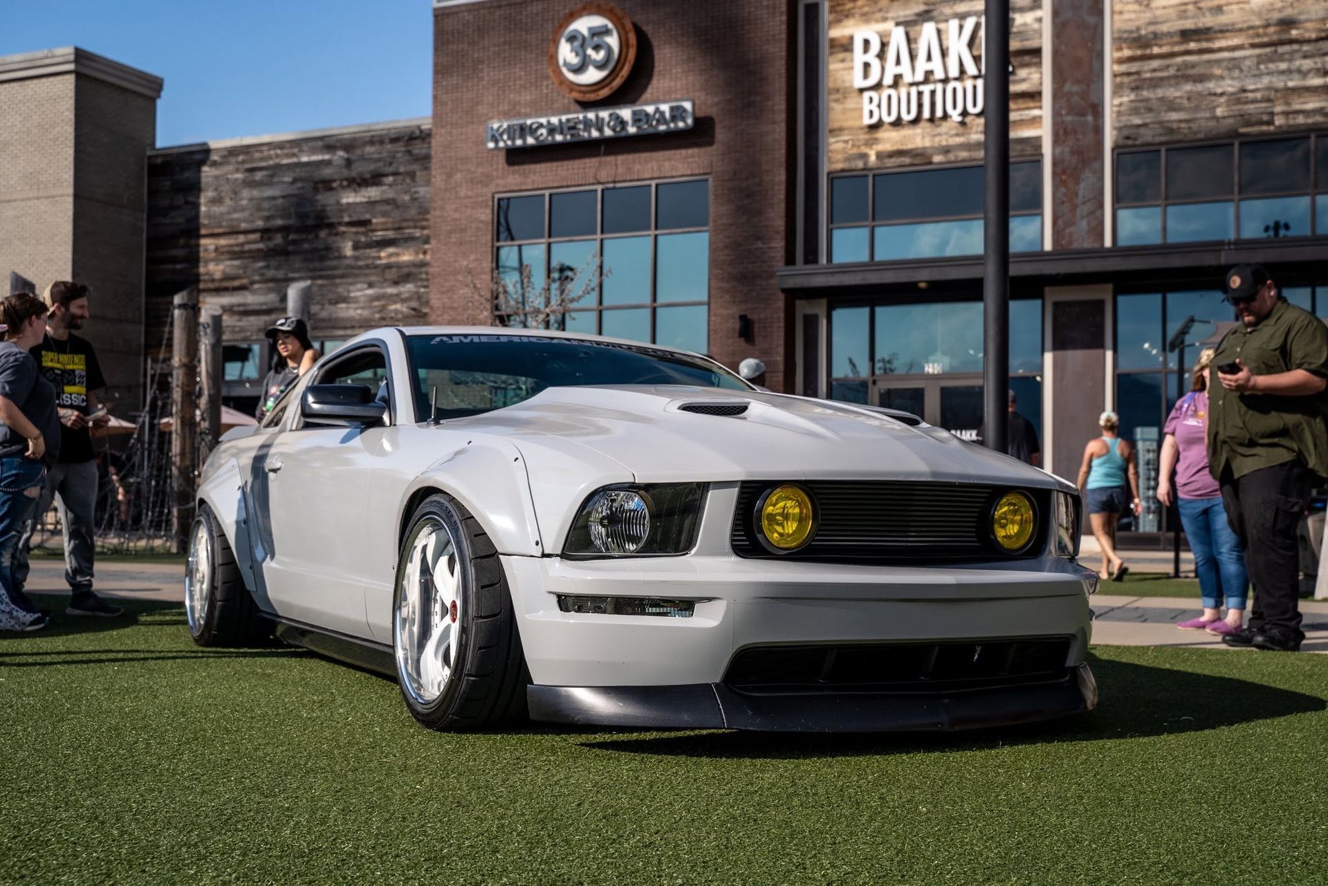 White classic muscle car on grass in front of a building, with people standing nearby