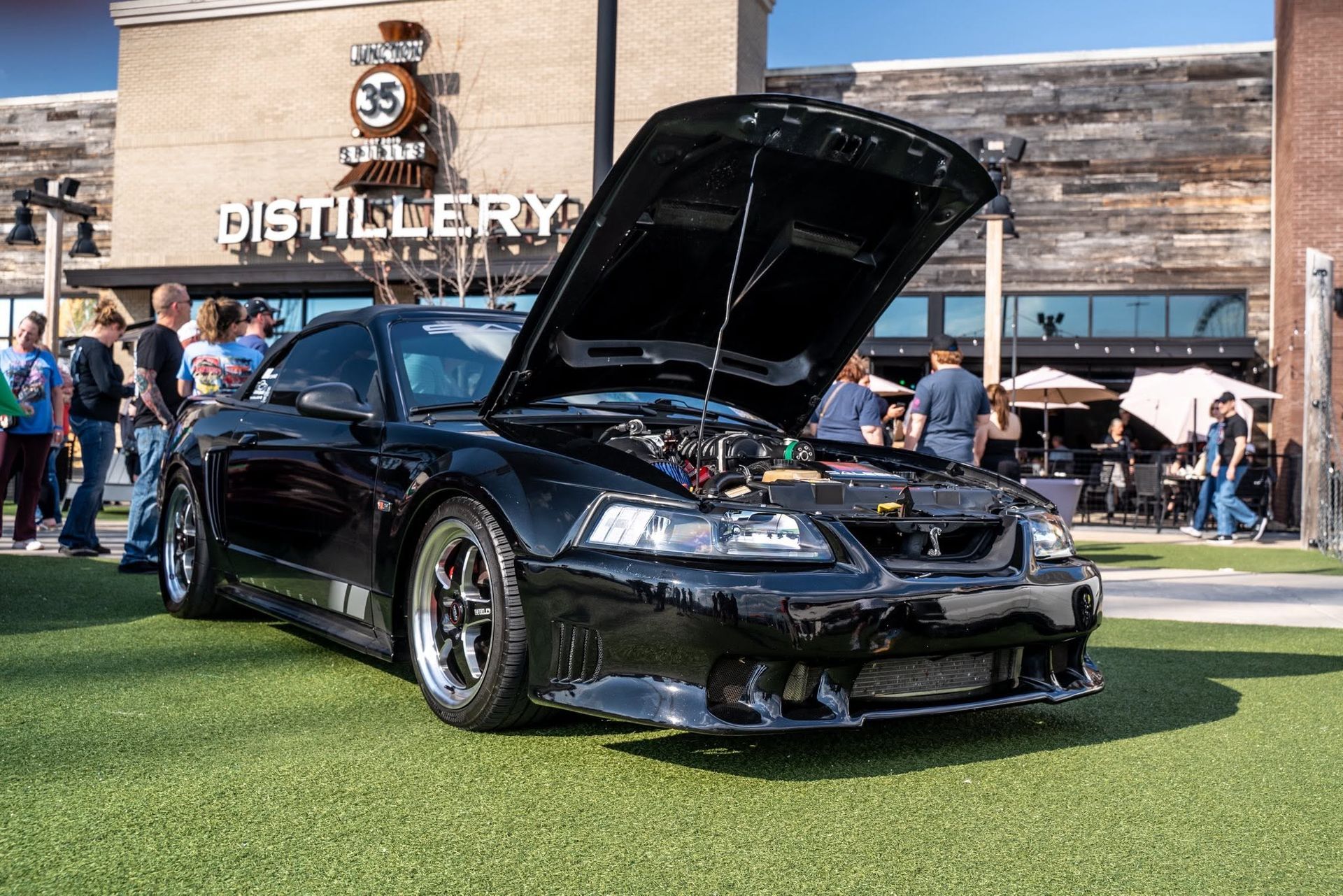 Black sports car with hood open at an outdoor car show in front of a distillery.