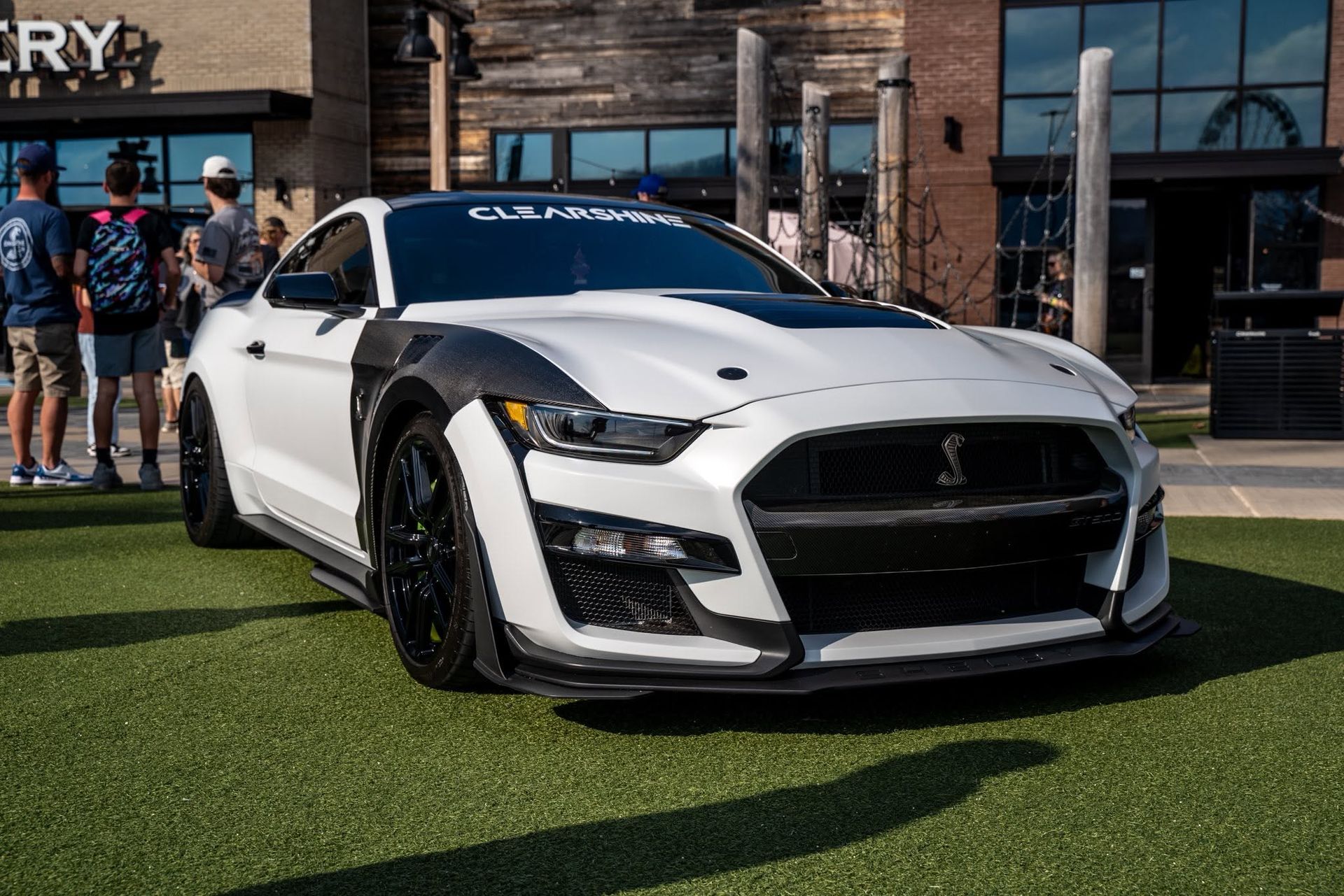White sports car on artificial turf outside a brick building, with people standing nearby