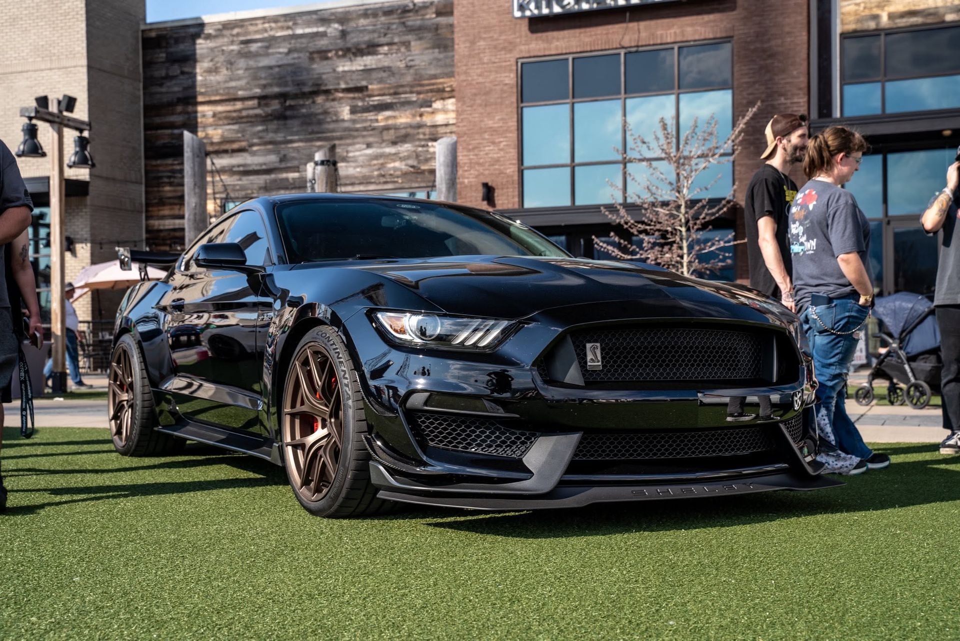 Black modified sports car on grass outside a brick building, with people walking nearby