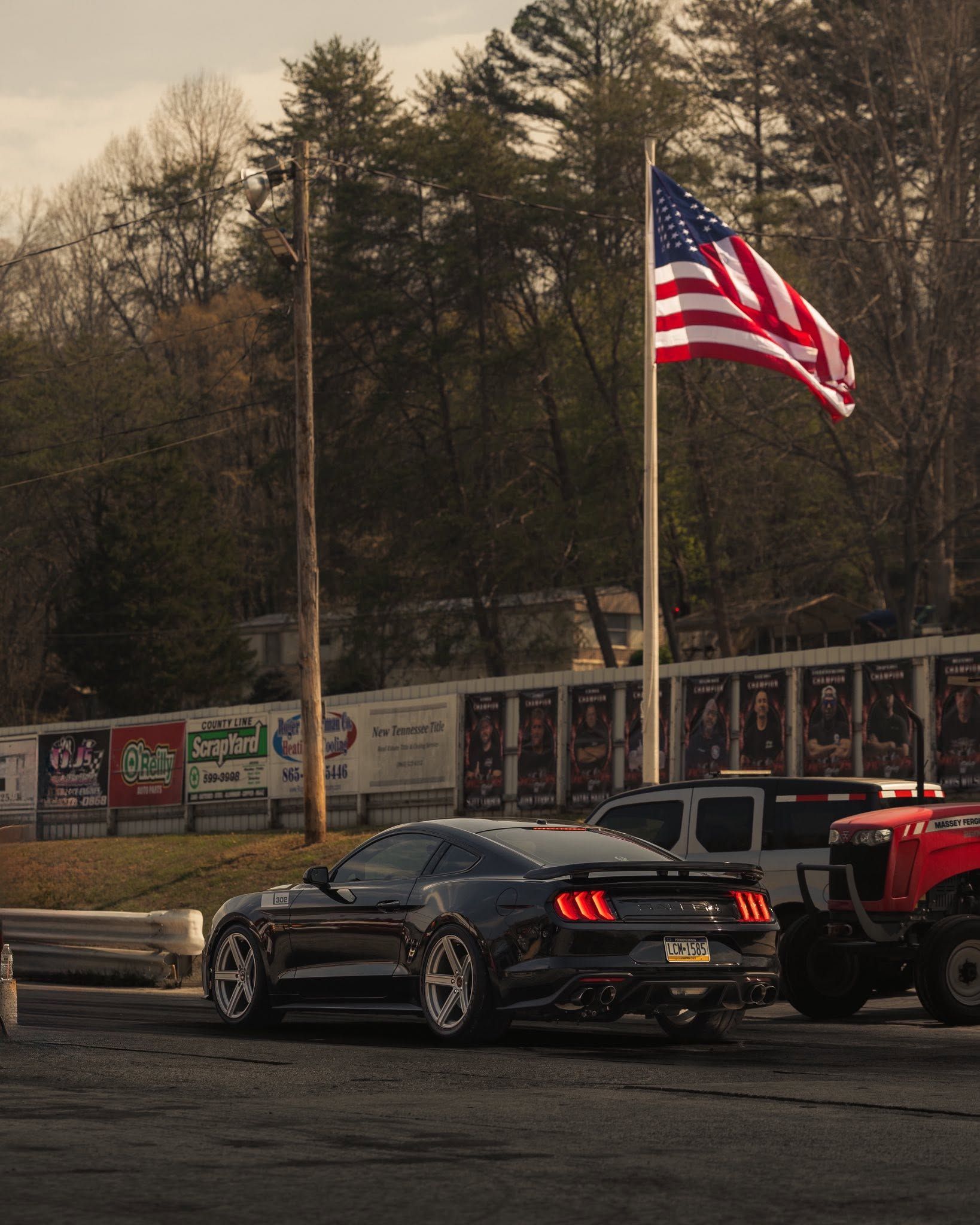 Black sports car near an American flag on a pole, with trees and a mural wall in the background