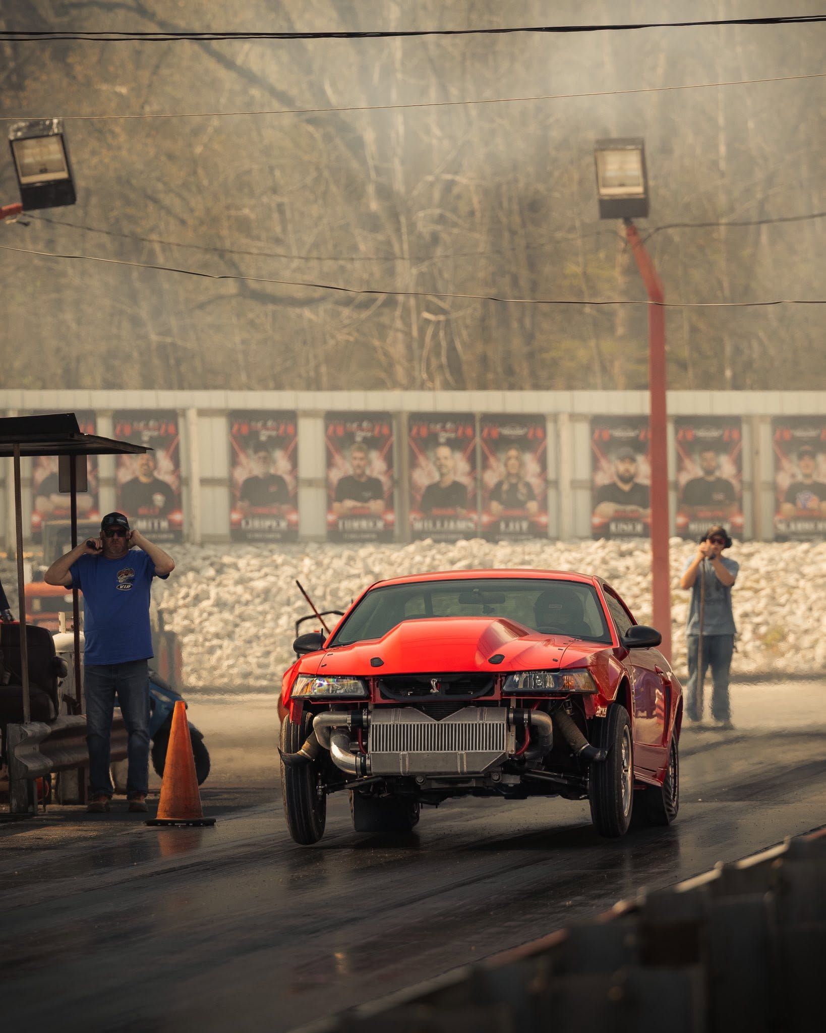 Red race car on a wet track with two people standing nearby under spray mist