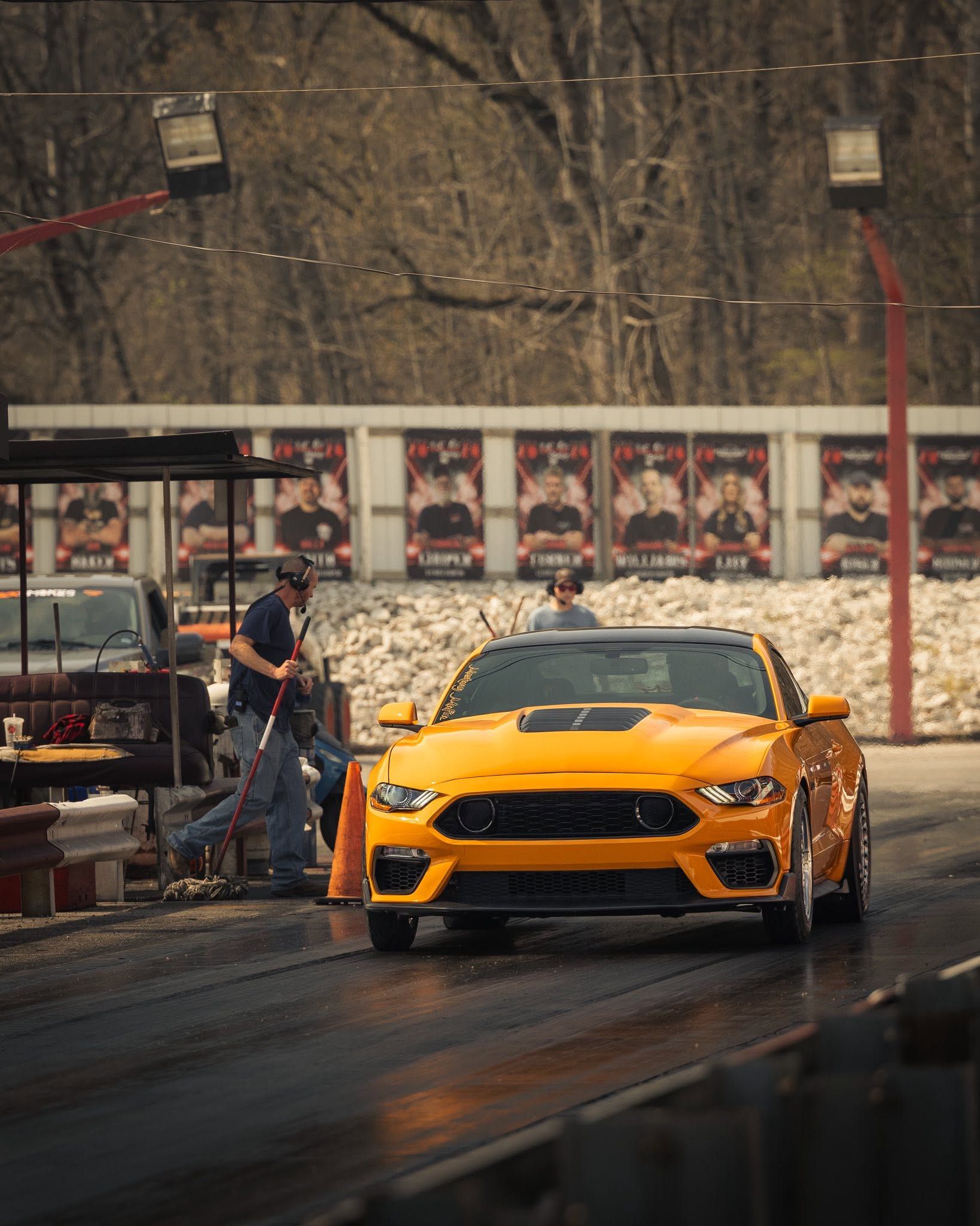 Yellow sports car on a wet road at a drag strip, with a person standing nearby.