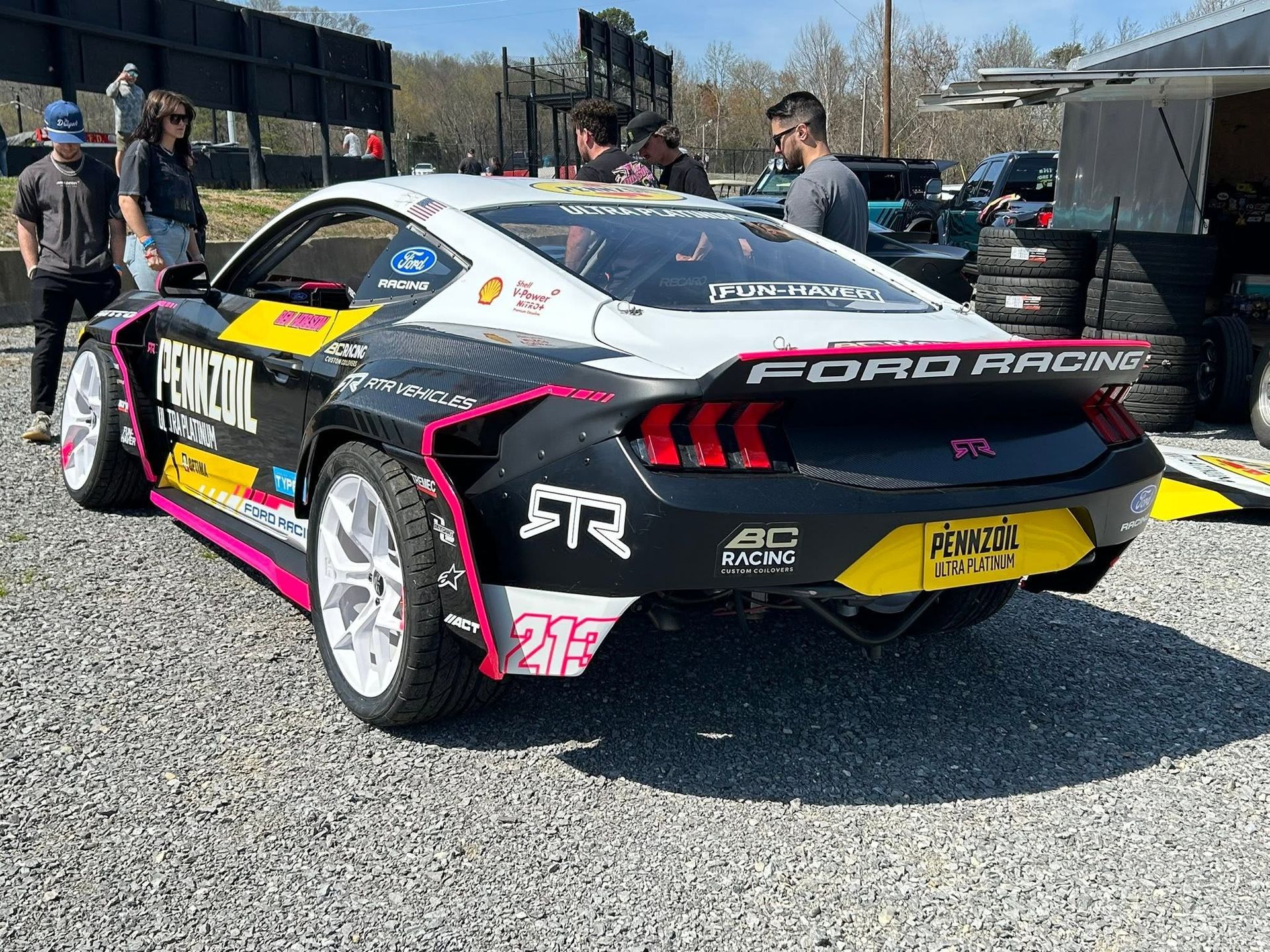 Ford race car with colorful livery parked on gravel, people standing around at an outdoor track.