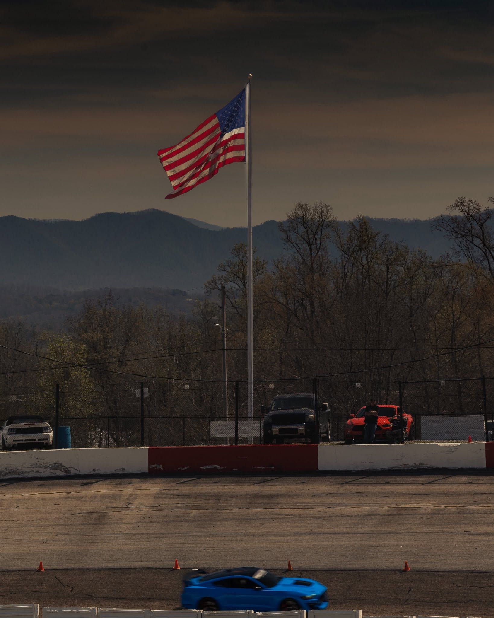 Race track with a blue car in front, an American flag waving above, and mountains in the background