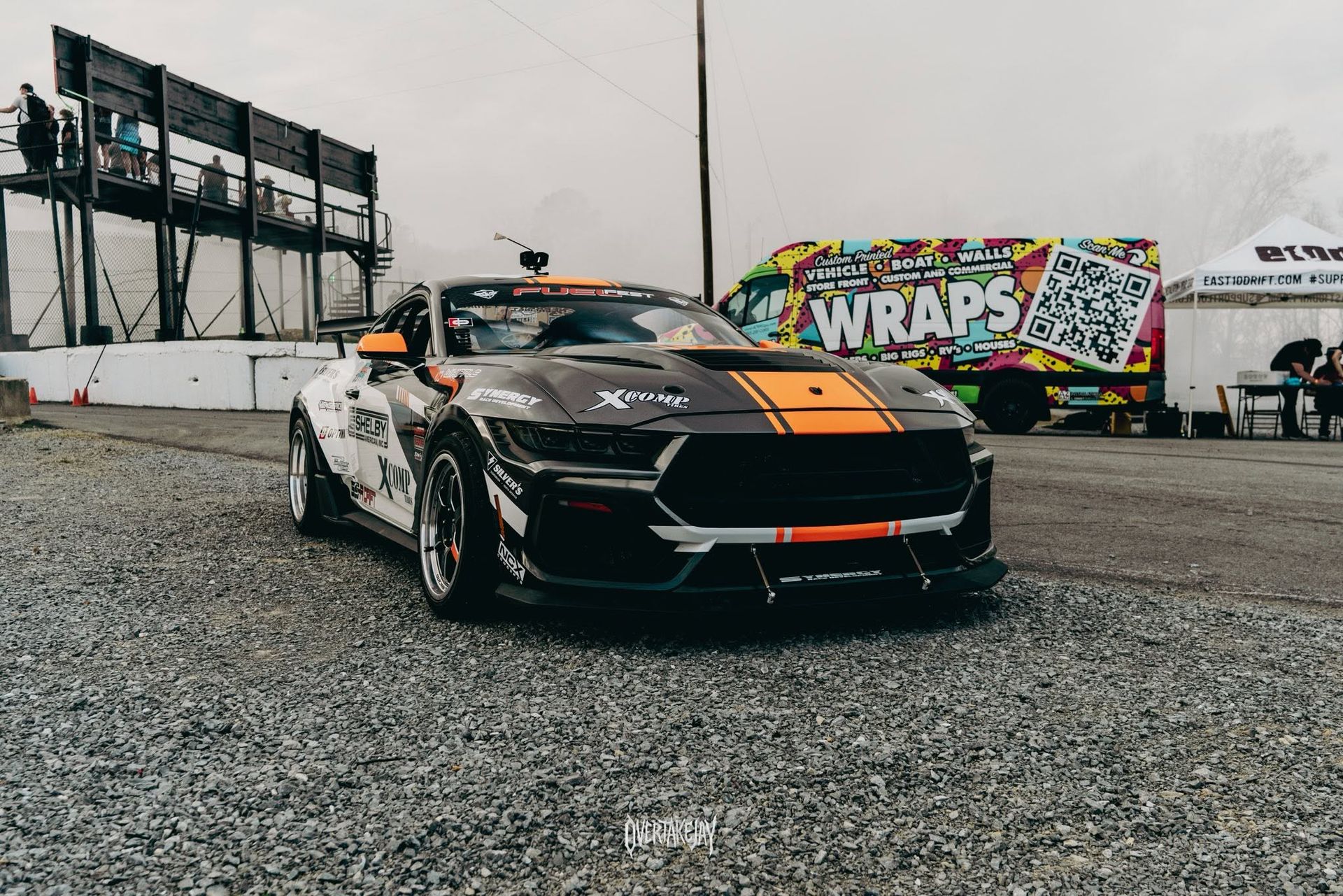 Black sports car with orange stripes parked on gravel near a colorful wraps banner and race track signage.