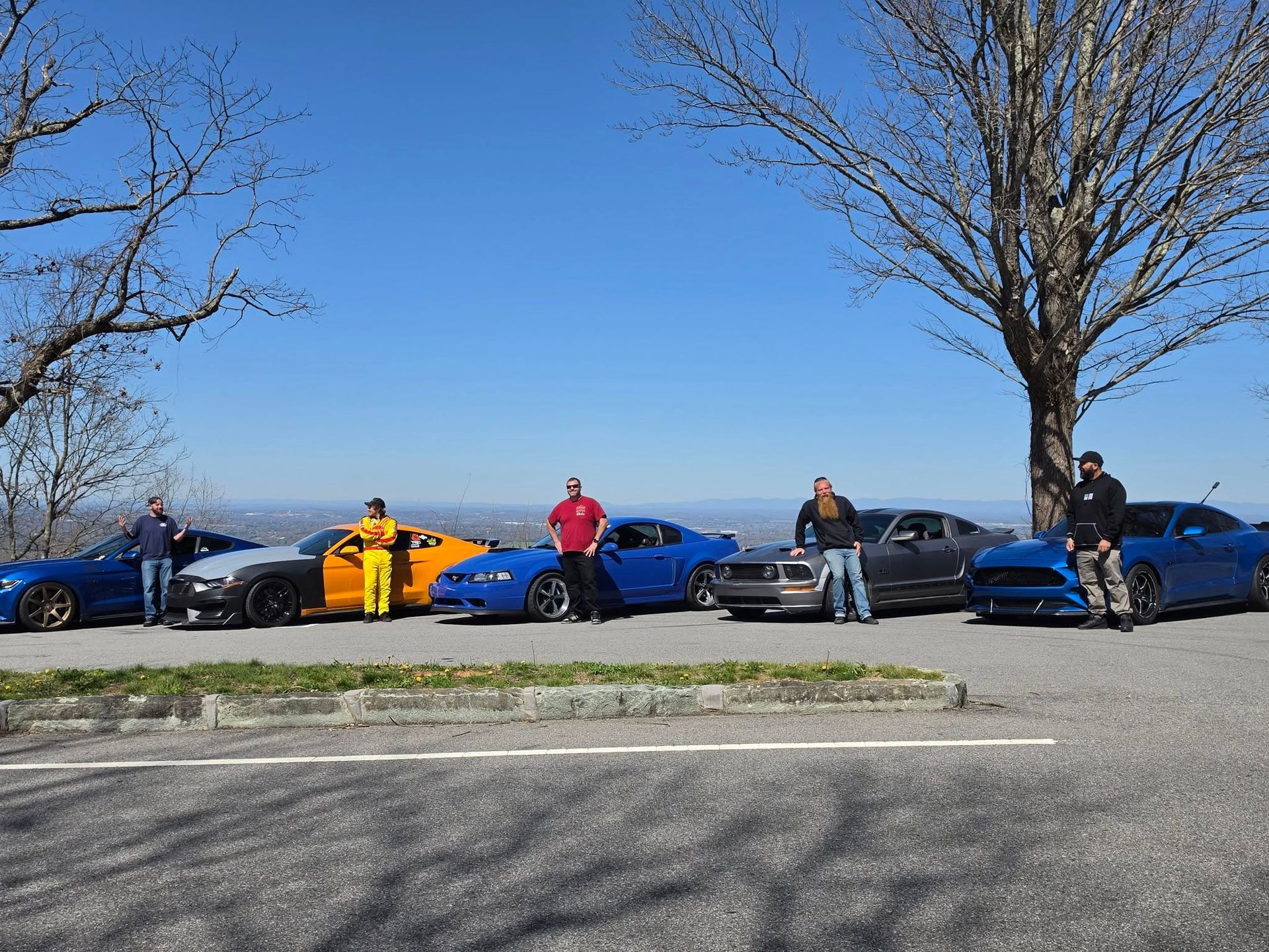Five people standing by colorful sports cars along a roadside under a clear blue sky