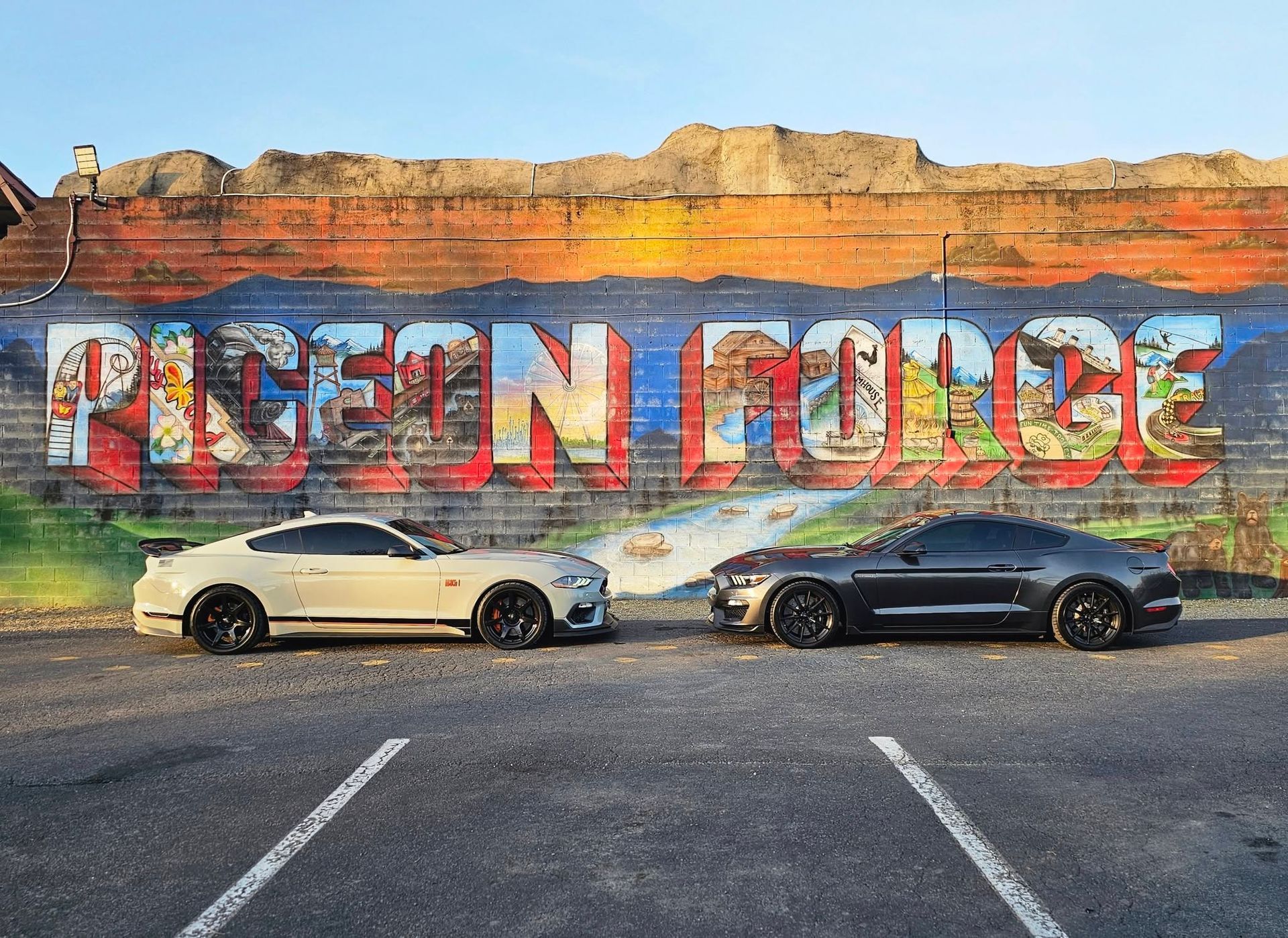 Two sports cars parked before a colorful cliff mural reading “PFEIFFER BIG SUR”