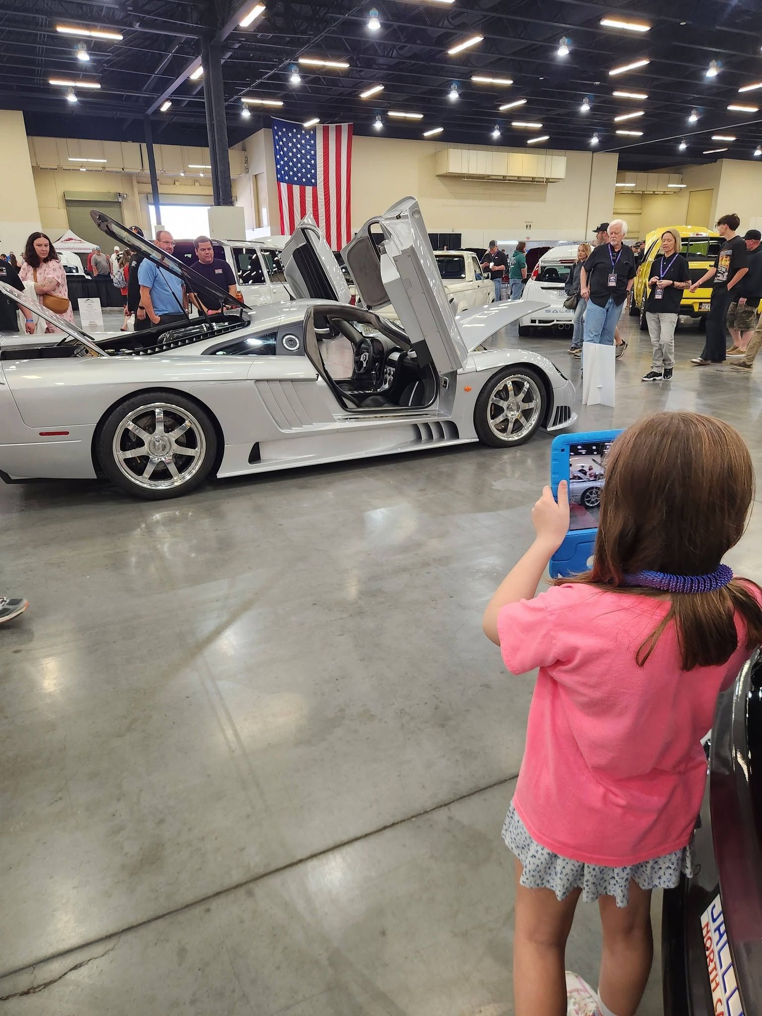 Silver sports car with scissor doors open at an indoor car show, people gathered around.