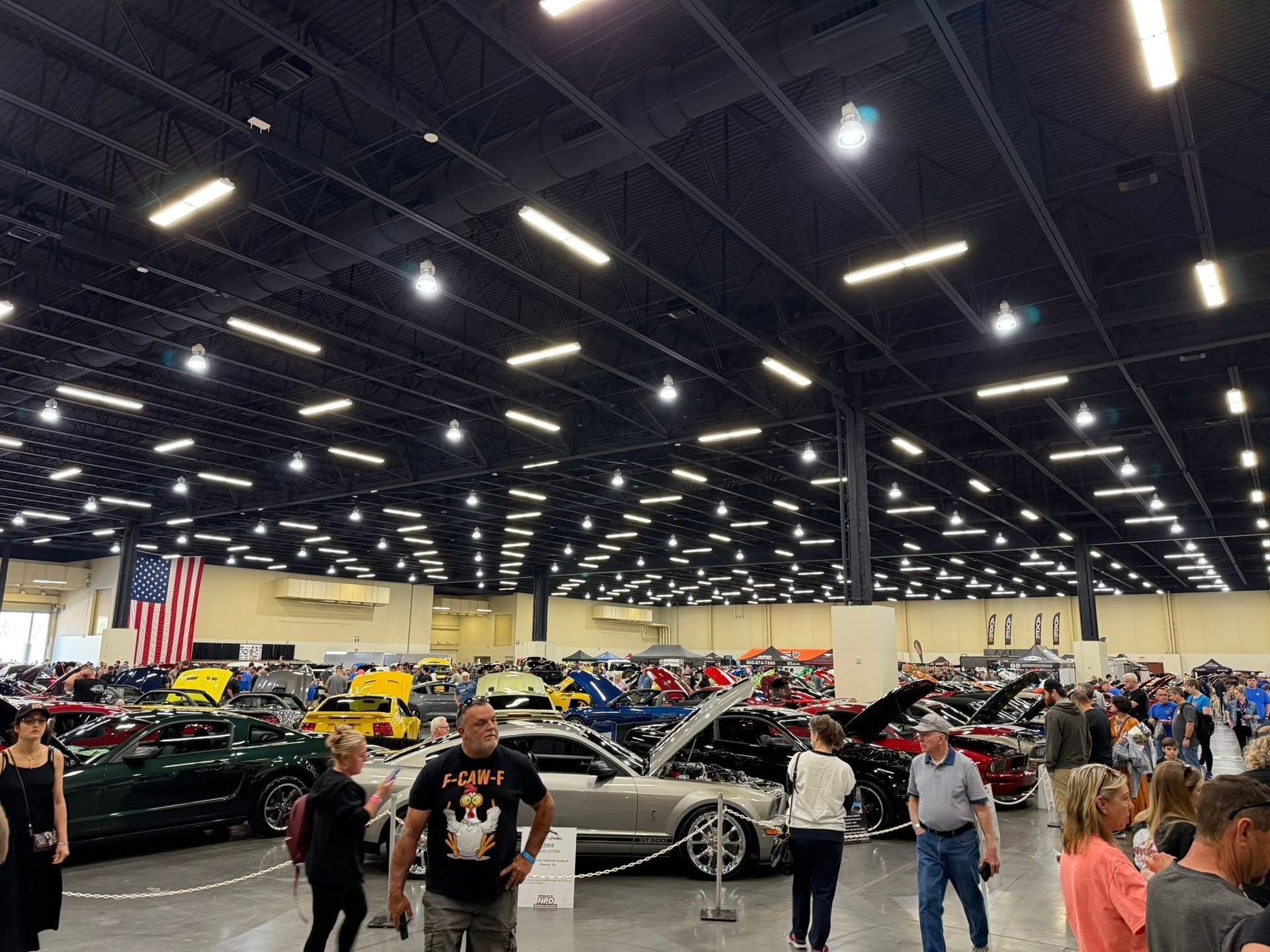 Indoor car show with rows of classic and modified cars under bright ceiling lights, people browsing around them