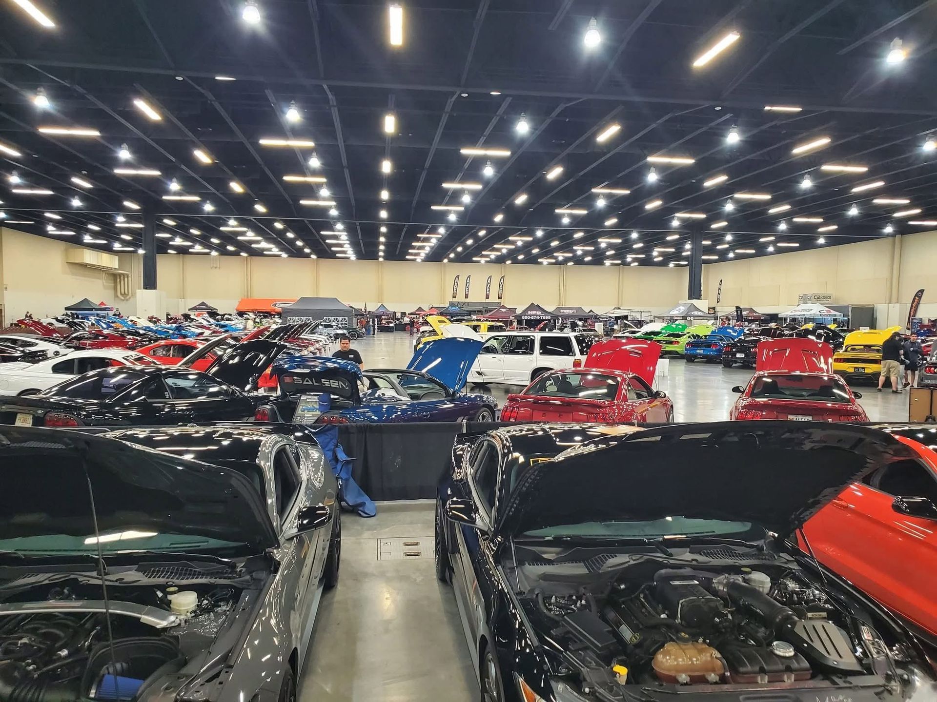 Warehouse full of parked cars under bright overhead lights, with several open hoods in the foreground
