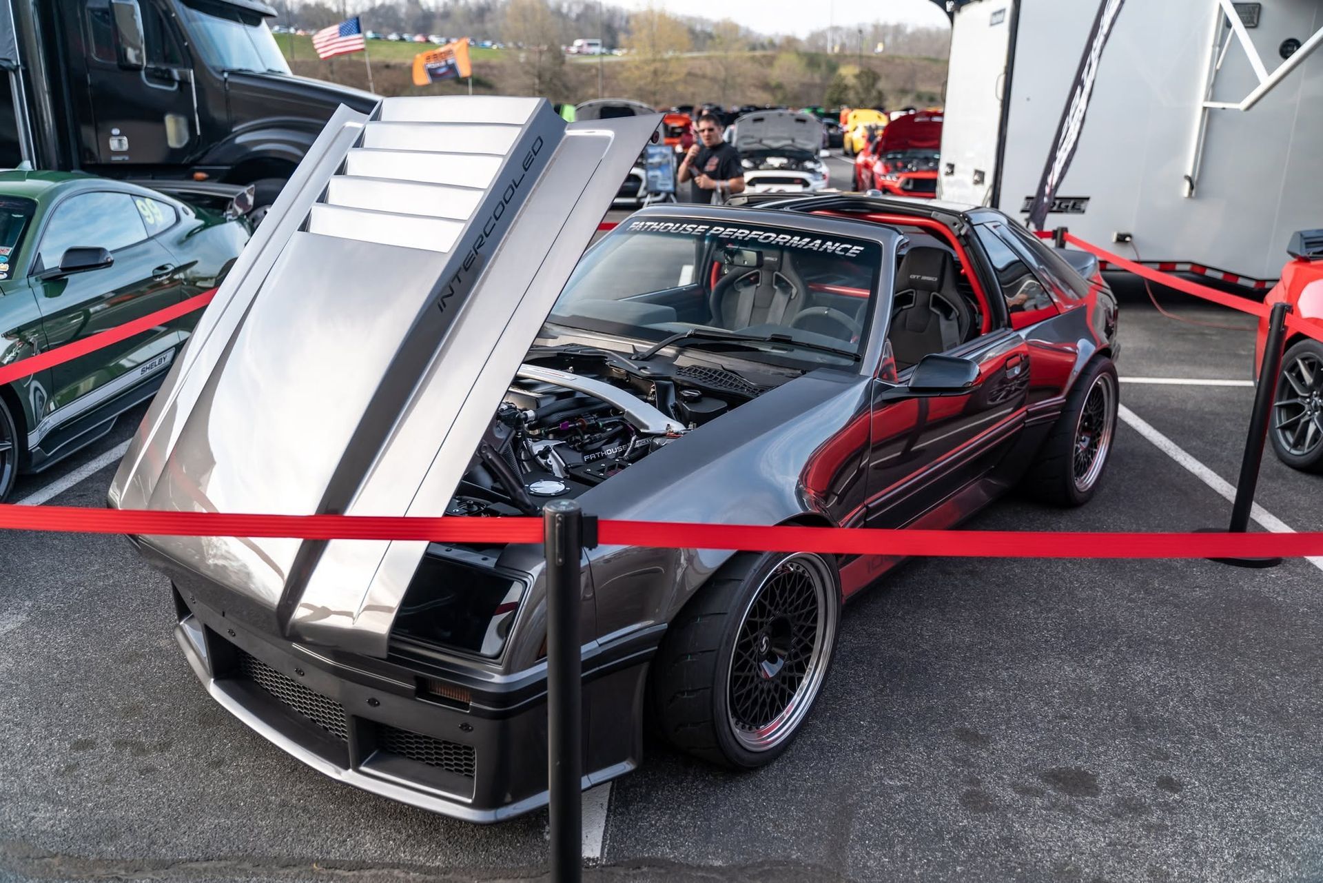 Black sports car with a raised hood at an outdoor car show, viewed behind a red rope barrier