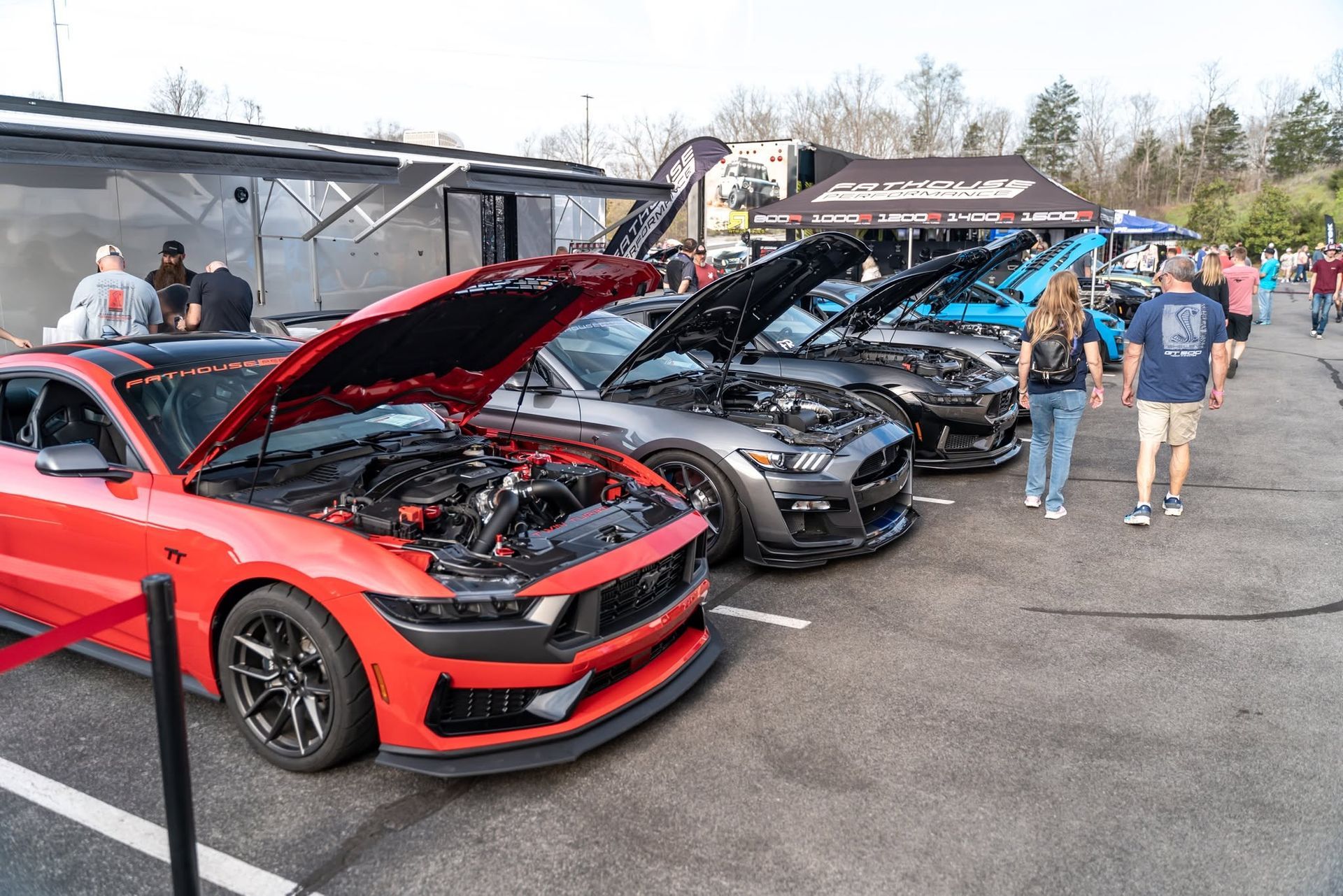Rows of sporty cars with hoods open at an outdoor car show, including a red car in the foreground and people walking nearby