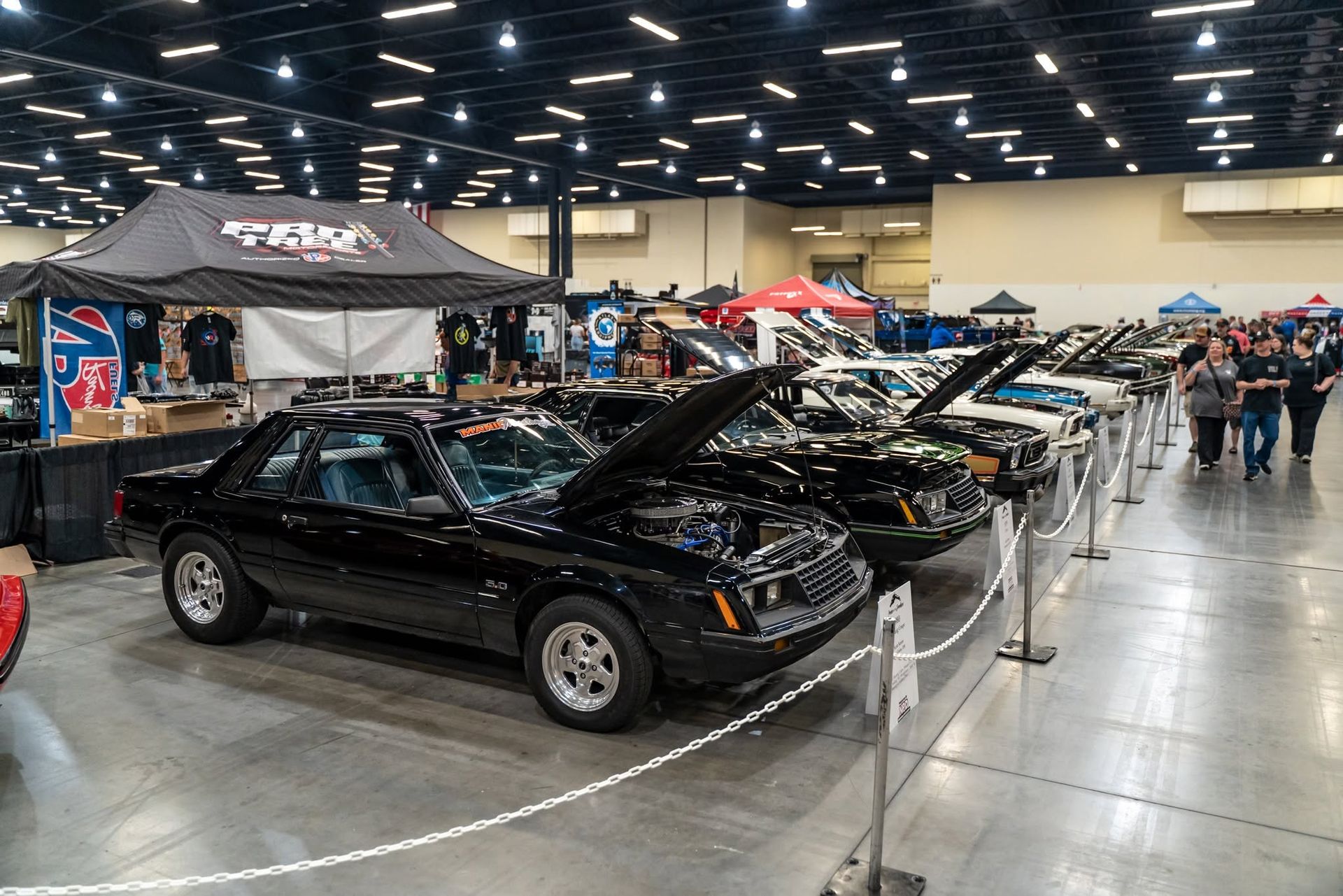 Black classic car at an indoor auto show, with rows of cars and vendor tents in a convention hall