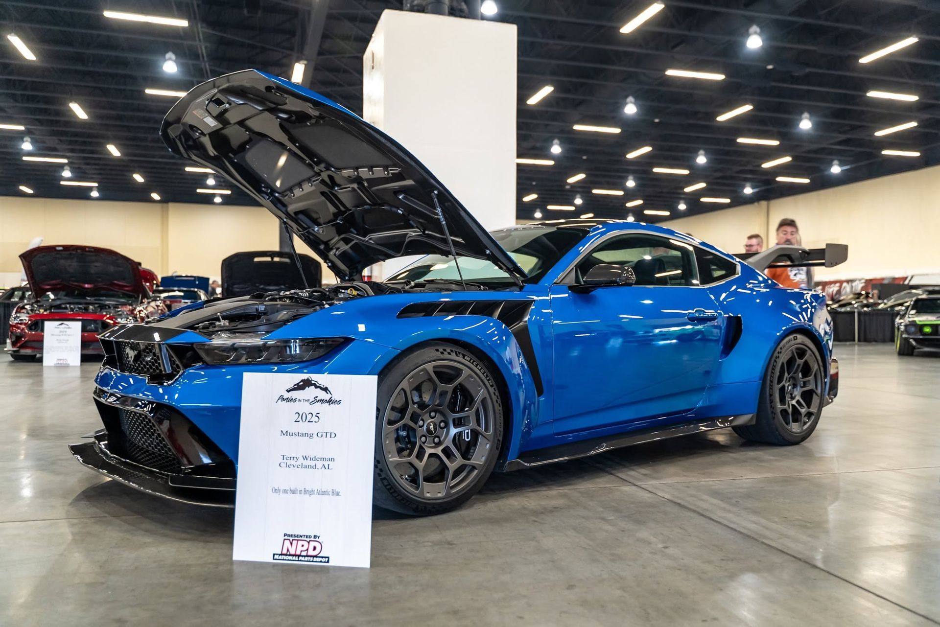 Blue sports car on display at an auto show with its hood open and a spec placard in front.