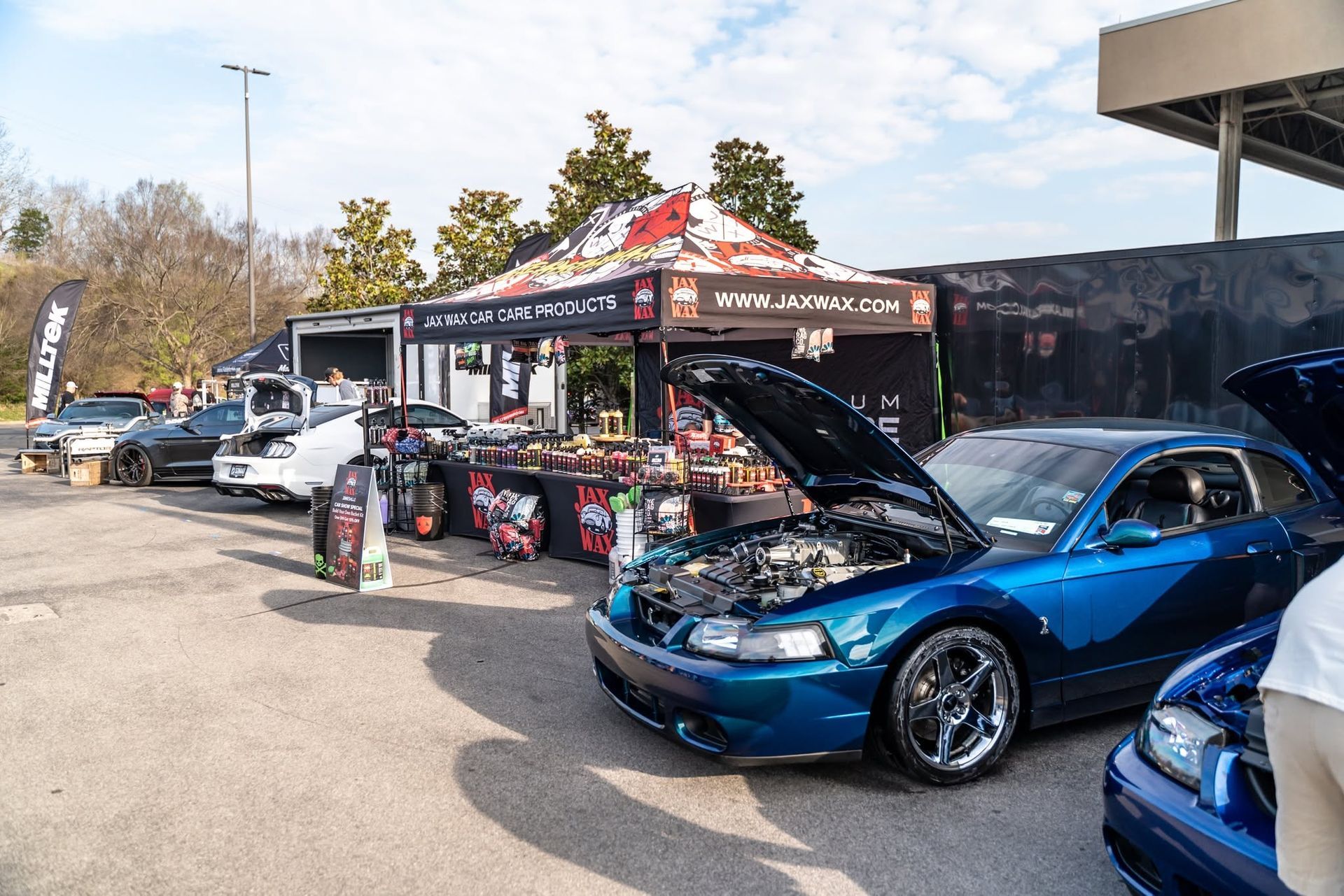 Blue car with open hood at an outdoor car show beside a vendor tent