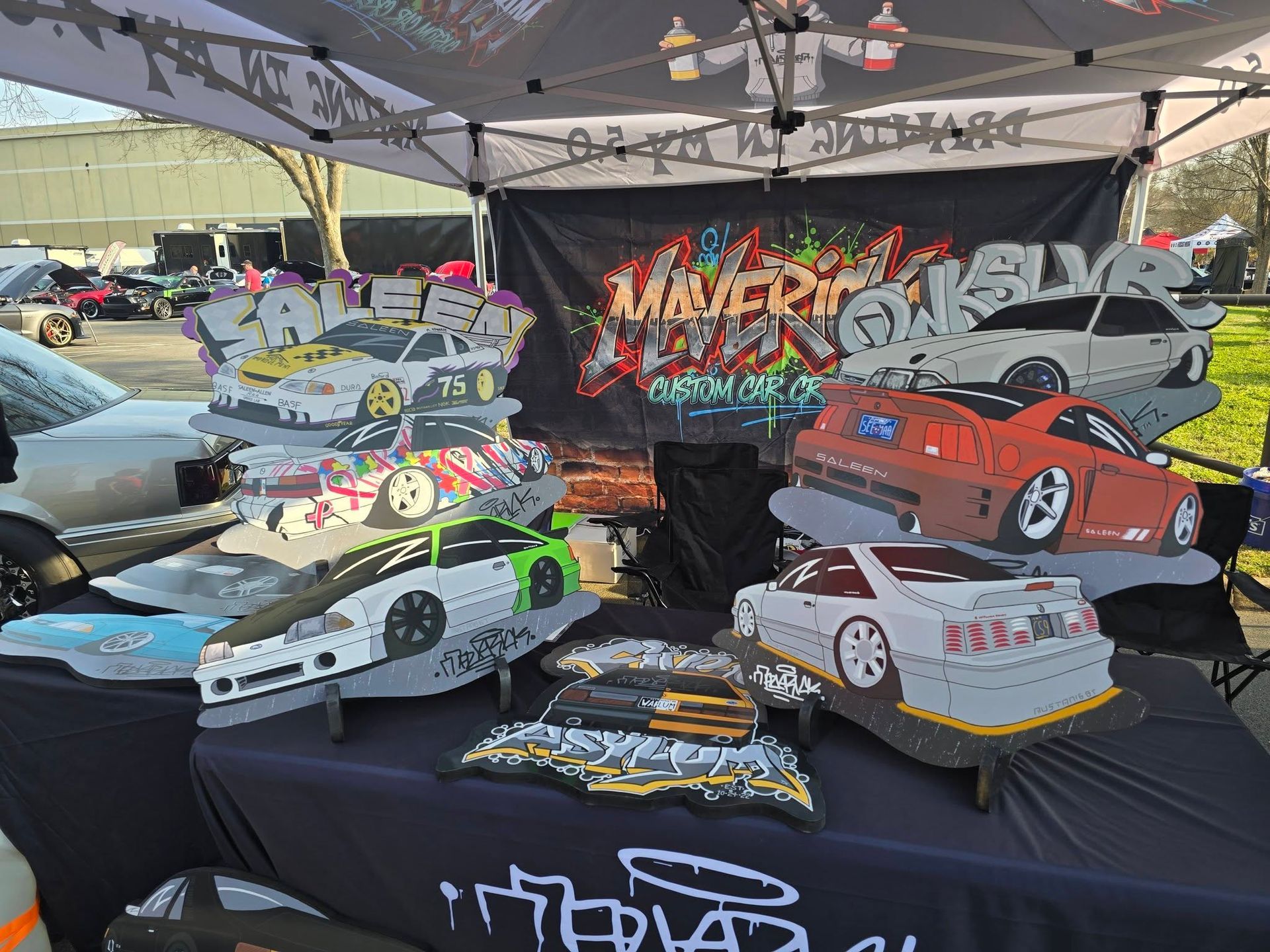 Colorful custom model cars displayed on a black table at an outdoor car show booth with graffiti backdrop