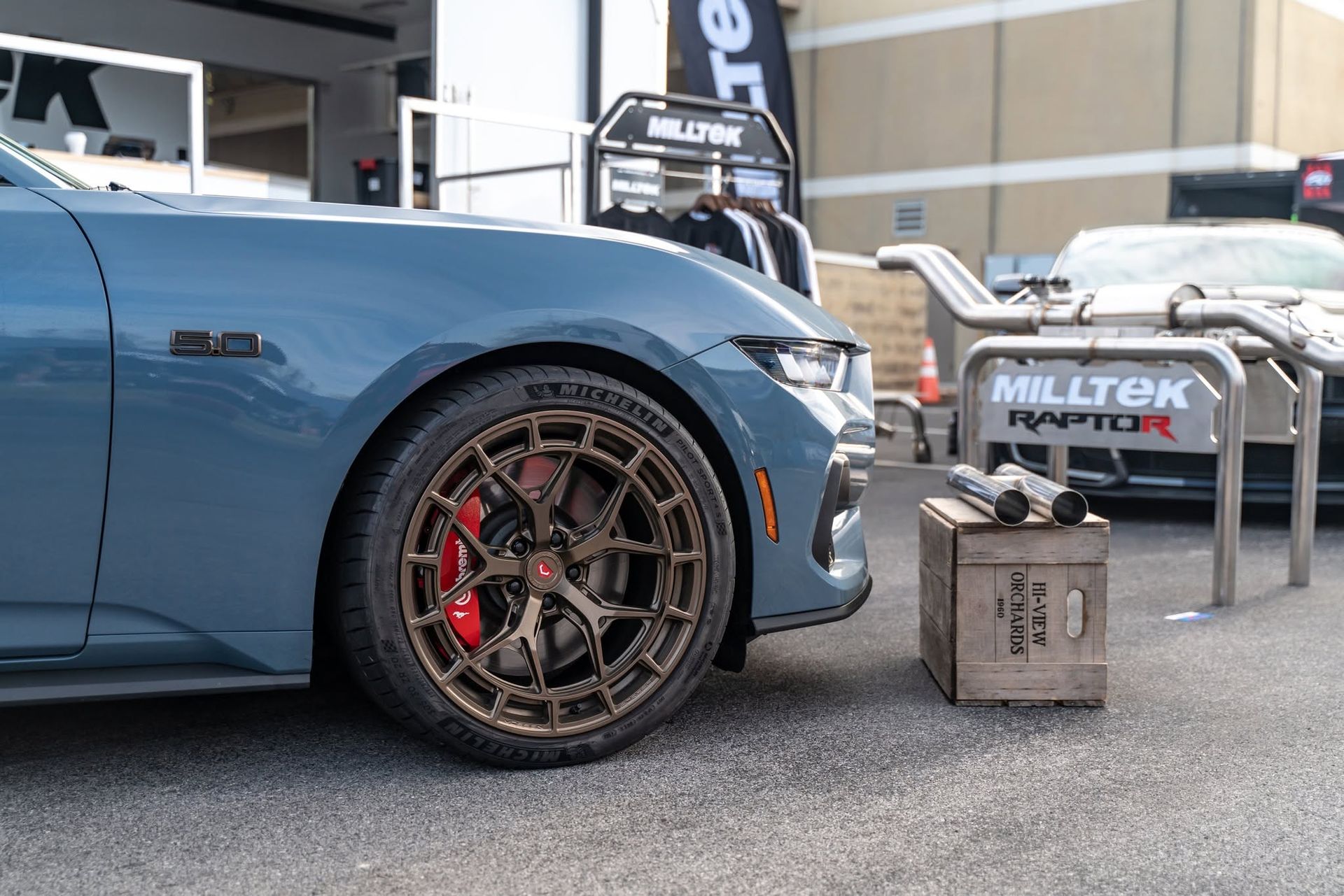 Blue sports car with bronze rims parked at an outdoor car show beside a small metal crate