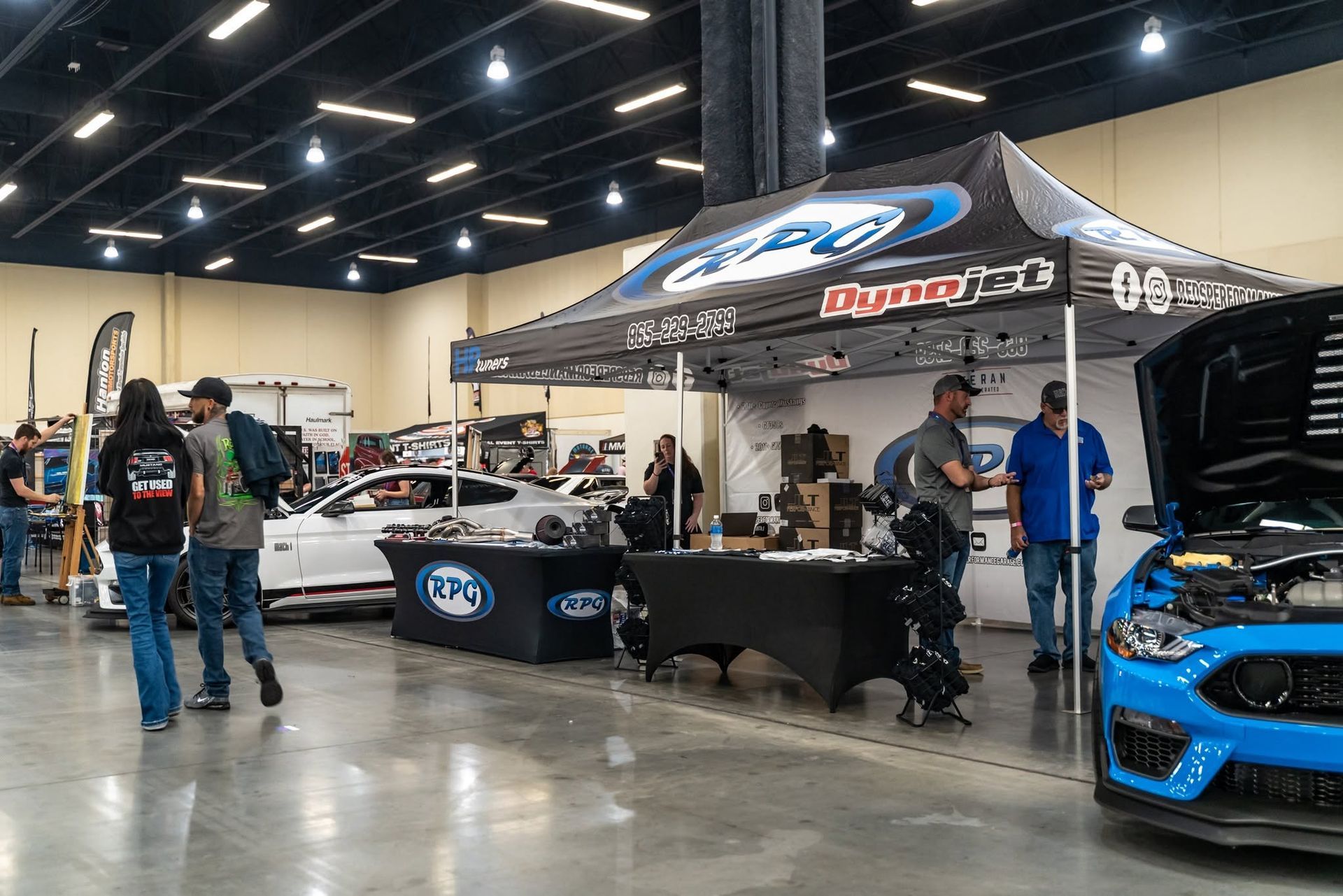 Indoor auto show with race car displays, a blue sports car, and people under branded tents