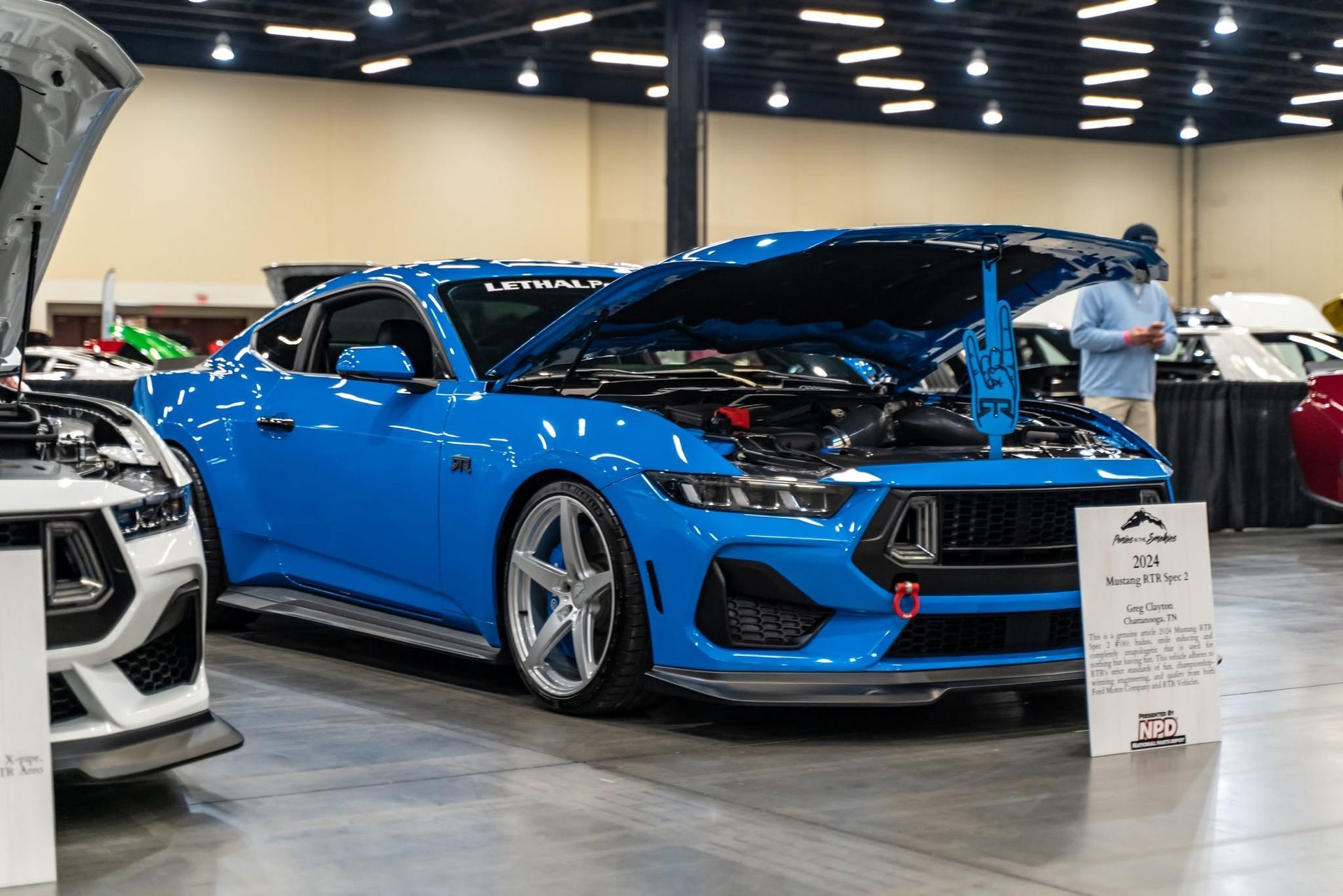 Blue sports car on display at an indoor auto show, with its hood open and a showroom sign in front.